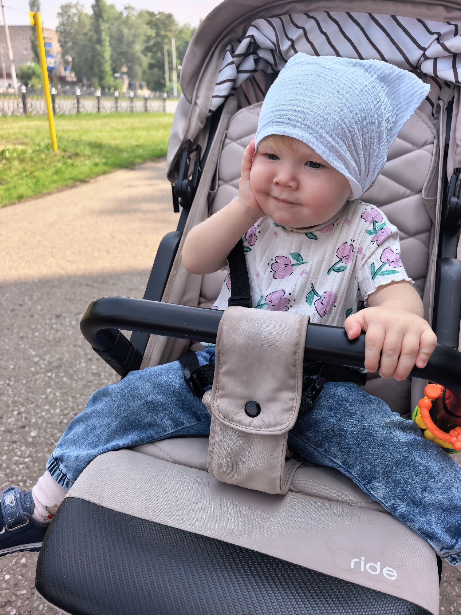A baby wearing a white headscarf and a white shirt with a floral pattern sits in a stroller. The baby has blonde hair and is resting their hand on their cheek. The stroller is beige and has a black handle. The background shows a paved path, green grass, and trees. A yellow pole and a fence are also visible in the background, suggesting an outdoor urban park setting. The scene appears to be in the city of Sterlitamak, Russia.