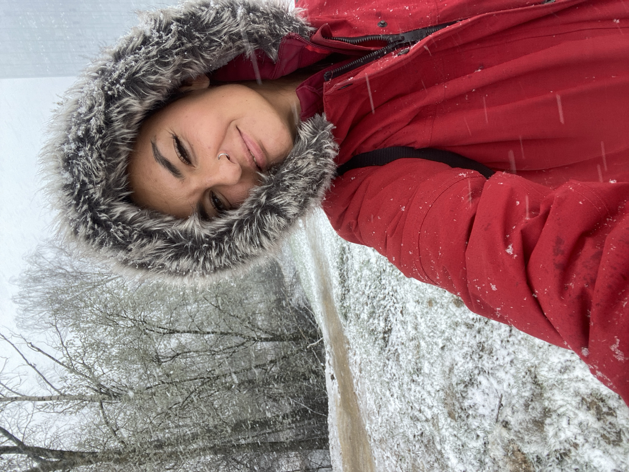 A woman in a red winter coat with a fur-trimmed hood takes a selfie in a snowy, wintry scene. The ground is covered in snow, and there are trees in the background, also dusted with snow. The location appears to be outdoors, likely a park or wooded area. The woman is facing the camera with a slight smile, wearing a nose ring. Snowflakes are visible in the air. The city of Khimki, Russia, cannot be confirmed from the image.