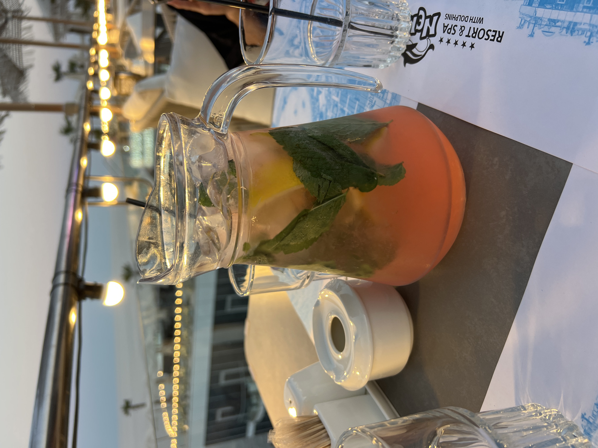 A pitcher of pink lemonade, garnished with mint leaves and lemon slices, sits on a table at a resort in Odesa, Ukraine. The pitcher is made of clear glass and has a handle. Next to the pitcher is a glass with ice and a black straw. In the background, there are blurred images of tables, chairs, and umbrellas, as well as string lights and the sky. The resort appears to be located near the sea. A paper menu with the text "RESORT & SPA WITH DOLPHINS" is also visible on the table.