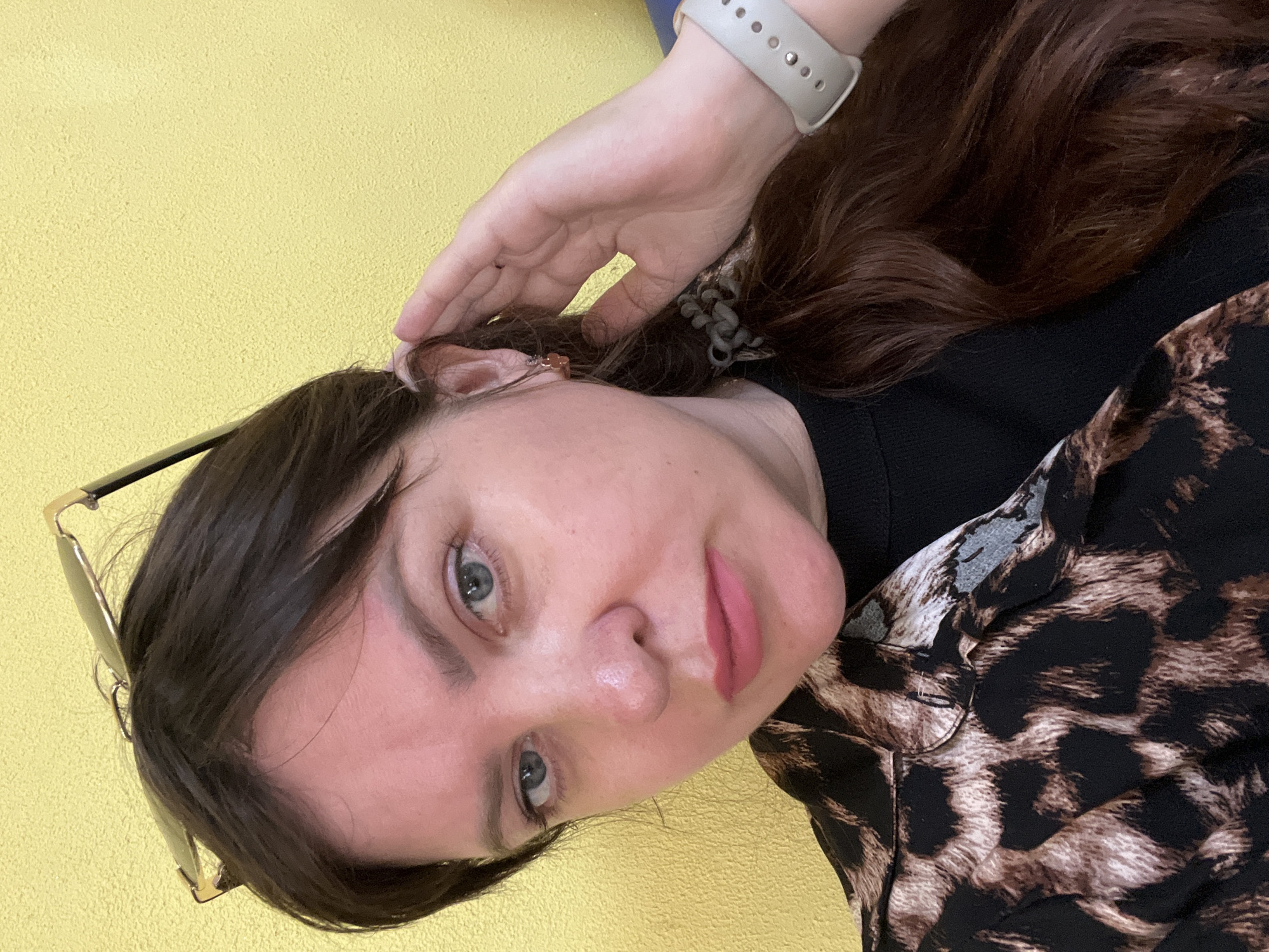 The image is a close-up selfie of a woman taken in portrait orientation, with her head tilted. She has brown hair and is wearing a black top with a leopard print collar. Her hand is touching her hair, and she is wearing a light-colored smartwatch on her wrist. She is also wearing glasses, which are resting on her nose. The background is a textured yellow surface, possibly a wall. The lighting appears to be natural, suggesting it might be daytime. The woman's expression is neutral, and there is no visible text in the image. The scene appears to be indoors, given the artificial lighting implied by the image's brightness and the absence of natural environmental cues.