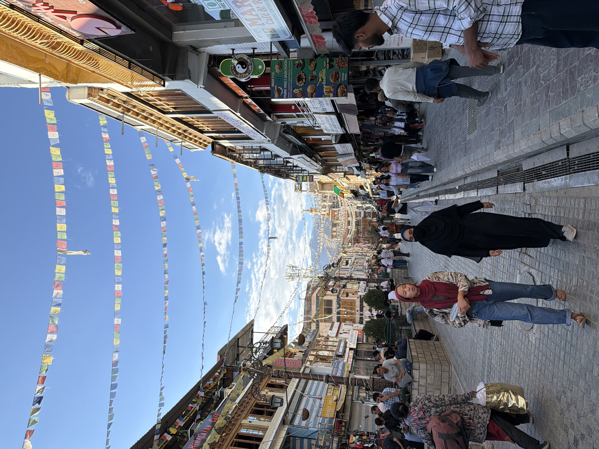 The image depicts a bustling street scene in Leh, India, likely a market area given the storefronts lining the street and the presence of pedestrians. Prayer flags are strung across the sky, a common sight in Buddhist regions like Leh. The architecture features multi-story buildings with traditional design elements and numerous shops. People are walking along the paved street, some dressed in traditional attire, while others are in casual Western clothing. There are several individuals seated on a low stone wall on the left side of the street, observing the activity. The overall atmosphere suggests a lively commercial district with cultural influences.