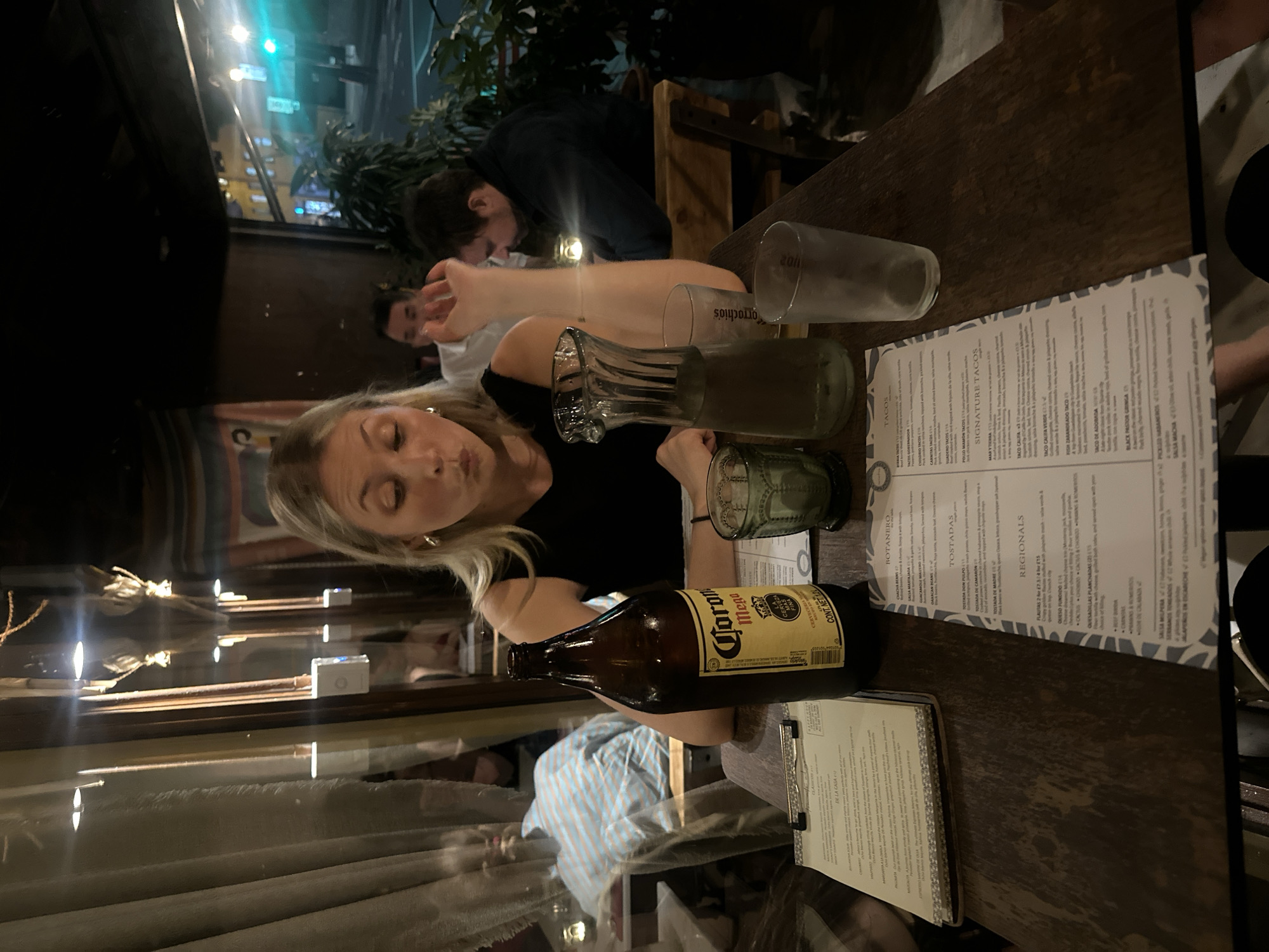 The image shows a table in a restaurant setting. On the table, there are several glasses, a bottle of Corona beer, and a menu. A woman with blonde hair is sitting at the table, and a man is visible in the background. The overall environment suggests an indoor dining space. The OCR reveals Spanish words and items typically associated with Mexican cuisine. It is not possible to determine the specific location in London from this image.