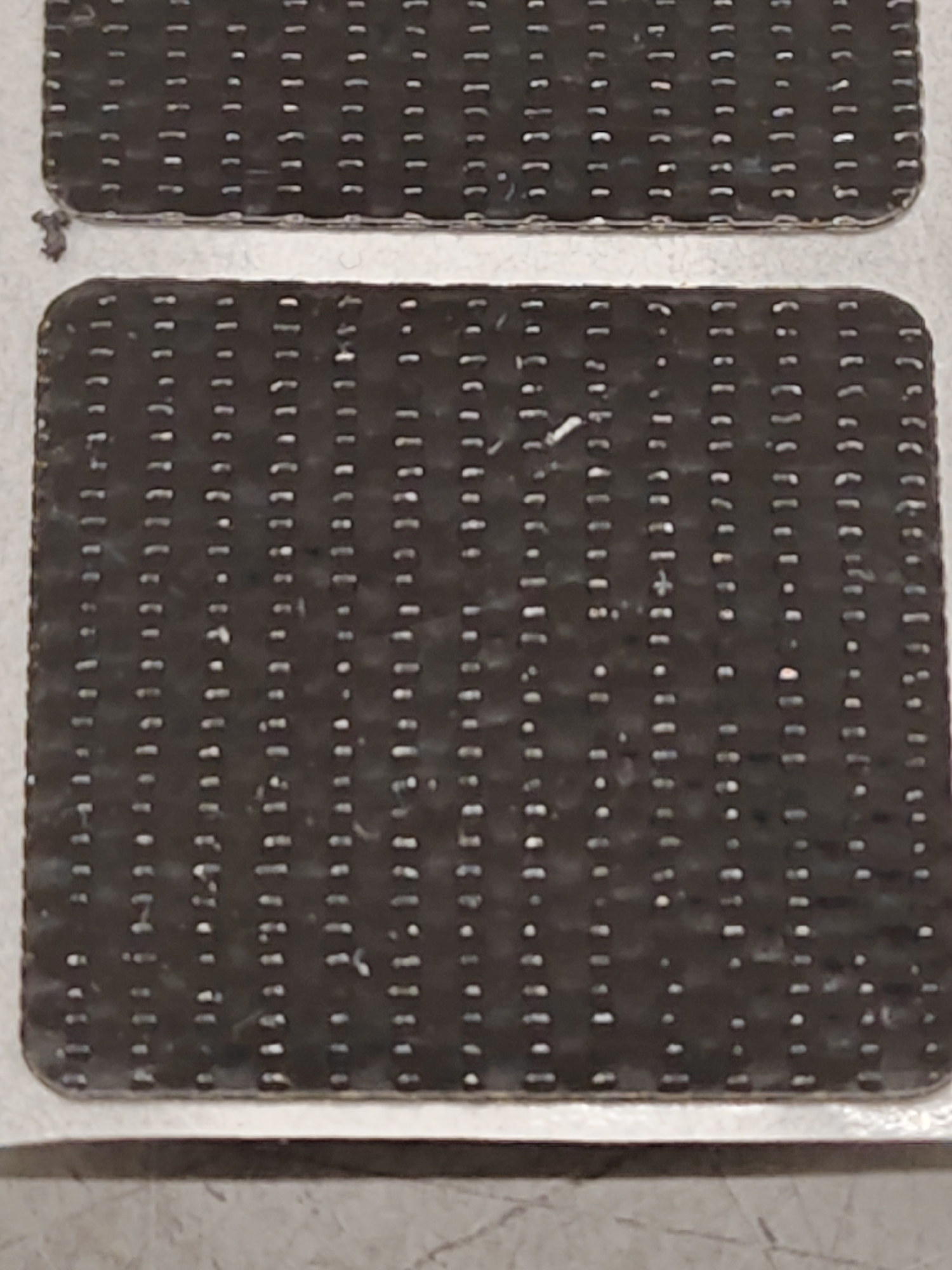 Two dark, rectangular objects with a patterned surface are shown in the image. The surface of each object is covered in a grid of small, raised dots or bumps. The top object appears to be slightly smaller and positioned above and to the left of the bottom object. The bottom object is larger and fills most of the frame. There are some white specks and smudges visible on the surface of the objects, as well as a small, dark, irregular object to the left of the top object. The background is a light-colored, textured surface.