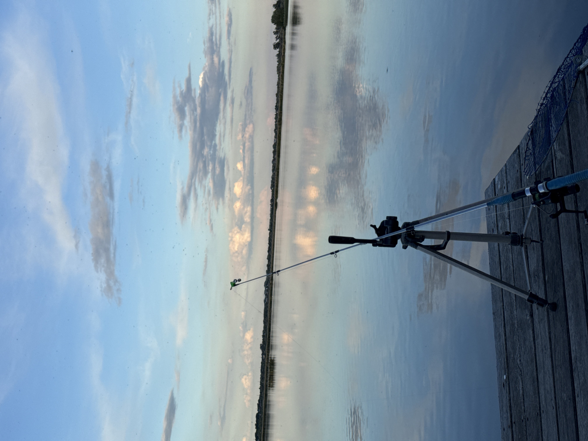 The image shows a fishing rod set up on a tripod, positioned on a wooden dock that extends into a body of water. A fishing net is also visible on the edge of the dock. The water reflects the sky, which is filled with clouds and light. The location is not specified.