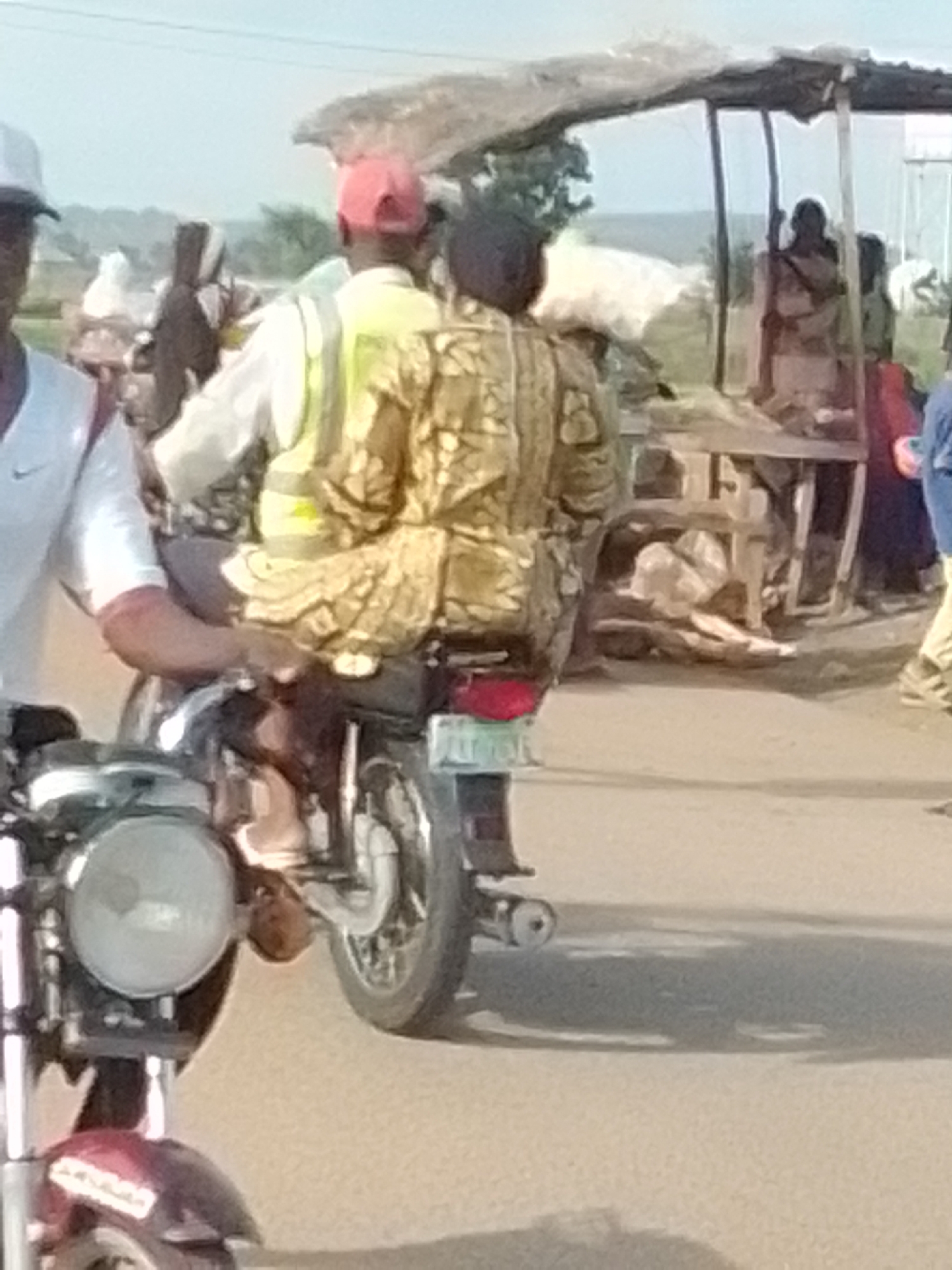 The image shows a street scene in Gombe, Nigeria. Two people are on a motorcycle. The person in the front is wearing a white shirt and a white cap. The person in the back is wearing a yellow and green reflective vest over a patterned shirt and a red cap. They are both looking in the same direction. In the background, there is a market stall with a thatched roof, and several people are gathered around it. The ground is paved, and there is a lot of activity in the area. The lighting suggests it is daytime, possibly late afternoon.