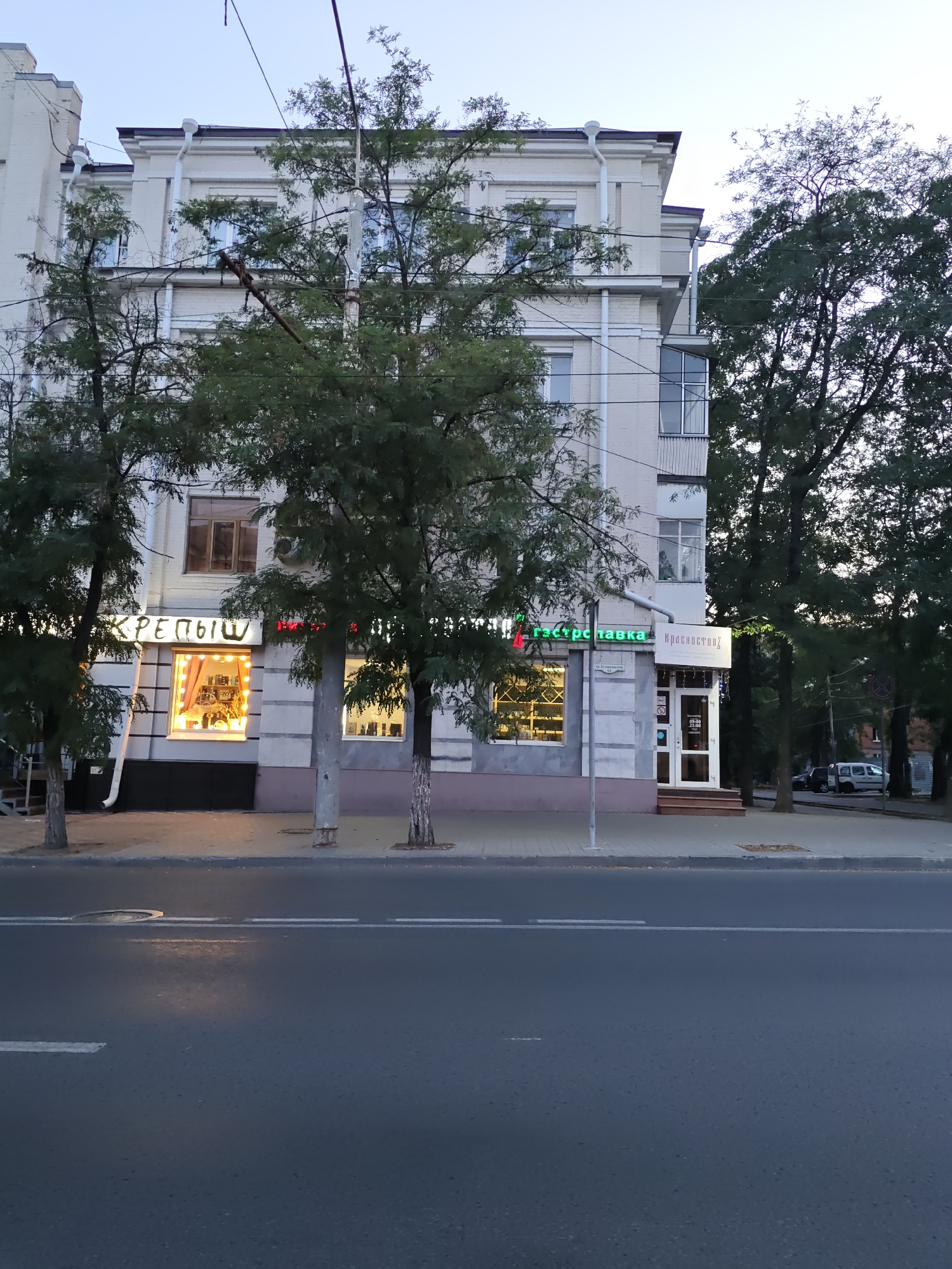 This is a photograph taken in Rostov-on-Don, Russia, depicting a street view in front of a building with commercial establishments. The building is a multi-story structure, likely residential or commercial, with a light-colored facade. In the foreground, a paved road with white lane markings is visible. The sidewalk in front of the building is also paved.
Several trees with green foliage are positioned along the sidewalk, partially obscuring the building. The building itself features several windows, some of which are illuminated. There are two visible storefronts with illuminated displays. The left storefront has large lettering that appears to be in Russian Cyrillic and displays various items inside. The right storefront, labeled "Красное Солнышко," is a shop entrance with a glass door and signage indicating operating hours.
Above the storefronts, there are more windows and architectural details of the building. Electrical wires are strung across the scene, typical of urban environments. In the background, more trees and parts of other buildings can be seen, along with a white van and other vehicles parked along the street. The sky is overcast and appears to be either early morning or late evening. No people are clearly visible in the image.