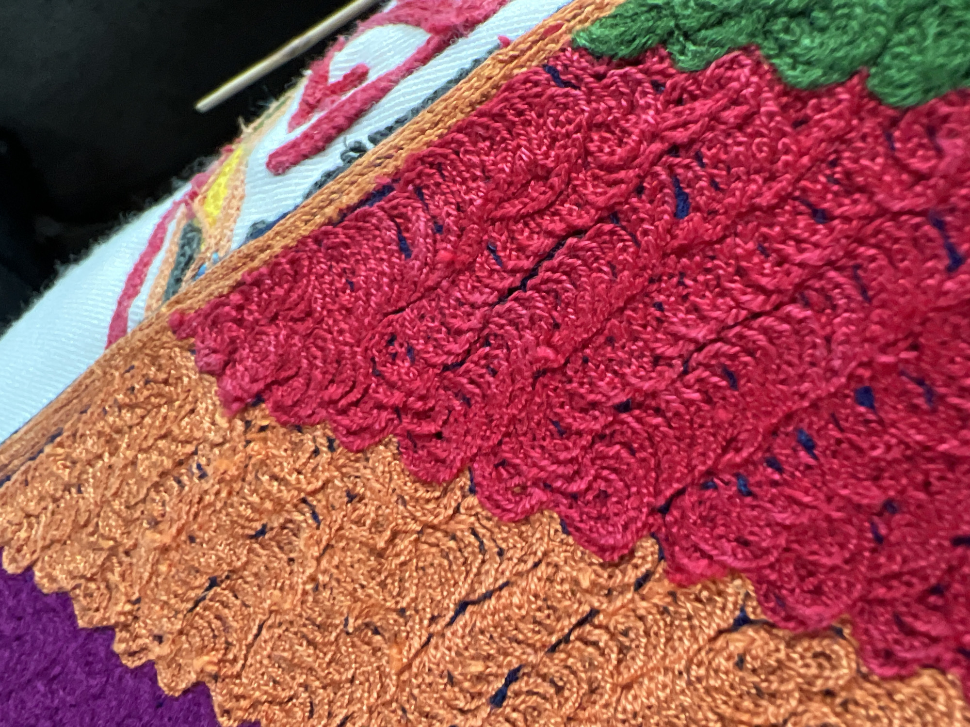 An extreme close-up depicts a section of vibrant, multi-textured embroidery or textile art, located in Slivovo, Bulgaria. The composition features distinct blocks of color with highly detailed stitching. A segment of deep purple fabric is visible on the upper left. Adjacent to it, a substantial area of orange stitching displays a dense, looped texture, creating a raised, intricate pattern with scattered dark blue threads. This orange section meets a prominent red section with a stepped, serrated edge. The red area exhibits an equally dense and textured stitch, potentially a complex chain stitch or knotted pattern, also interspersed with dark blue threads, forming a wave-like or scaled appearance. A bright green section, similarly textured, is partially visible at the bottom right, continuing the stepped pattern. On the upper right, a portion of white fabric with simpler embroidered lines in red and yellow is discernible. A slender, silver-toned needle or tool extends into the frame from the right, implying ongoing manual craftwork. The background behind the white fabric is dark and out of focus.