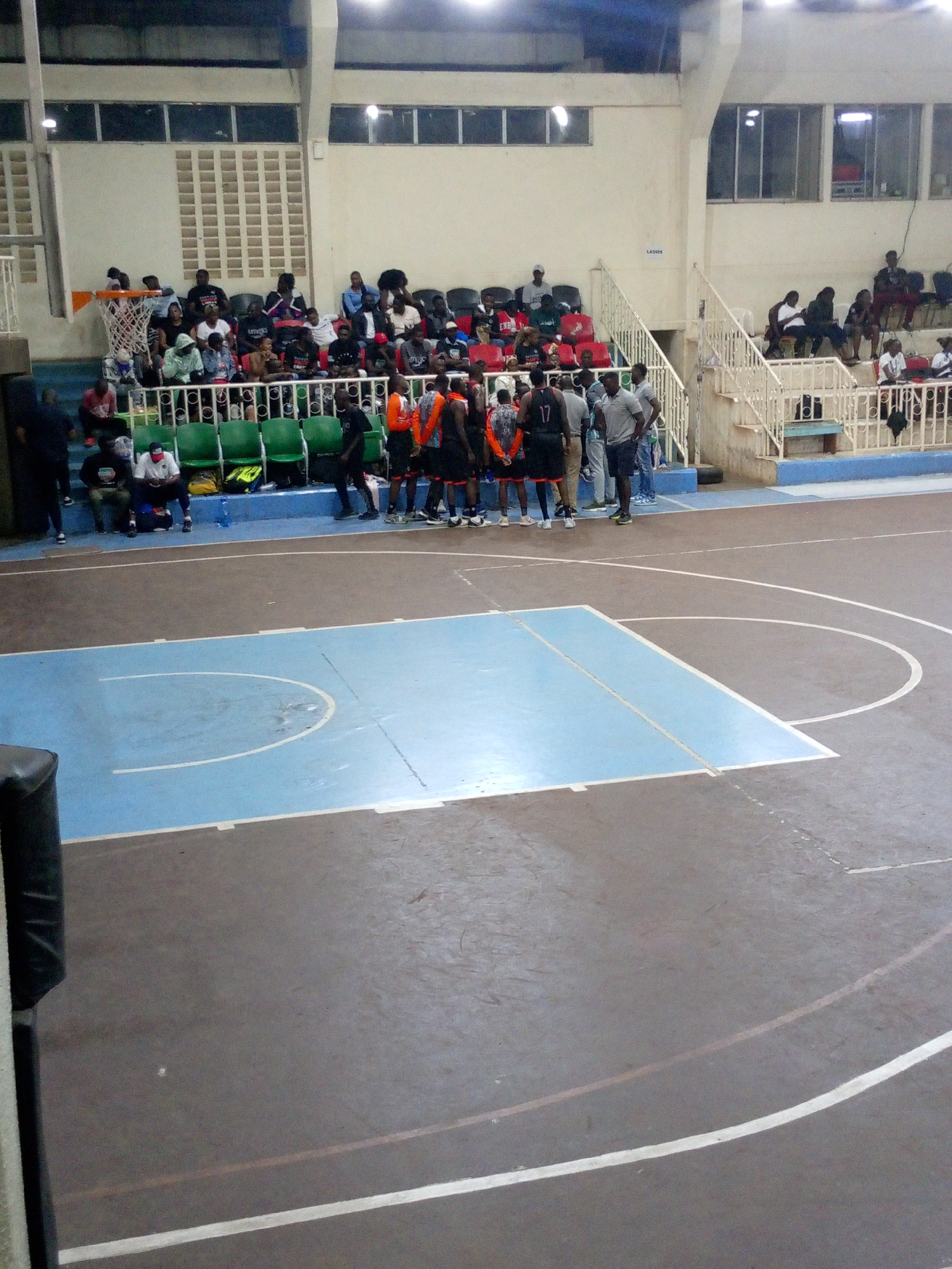 The image shows a basketball game or practice in progress at an indoor court. The location appears to be in Nairobi, Kenya. A group of players is huddled on the court near the free-throw line, with other players sitting on chairs along the sidelines. Spectators are seated in elevated seating areas on either side of the court. The court has a blue-painted area at each end, and white lines mark the boundaries and key areas. The overall lighting is somewhat dim, and the environment is a typical gymnasium setting. A sign that says "Ladies" can be seen above the seating area.