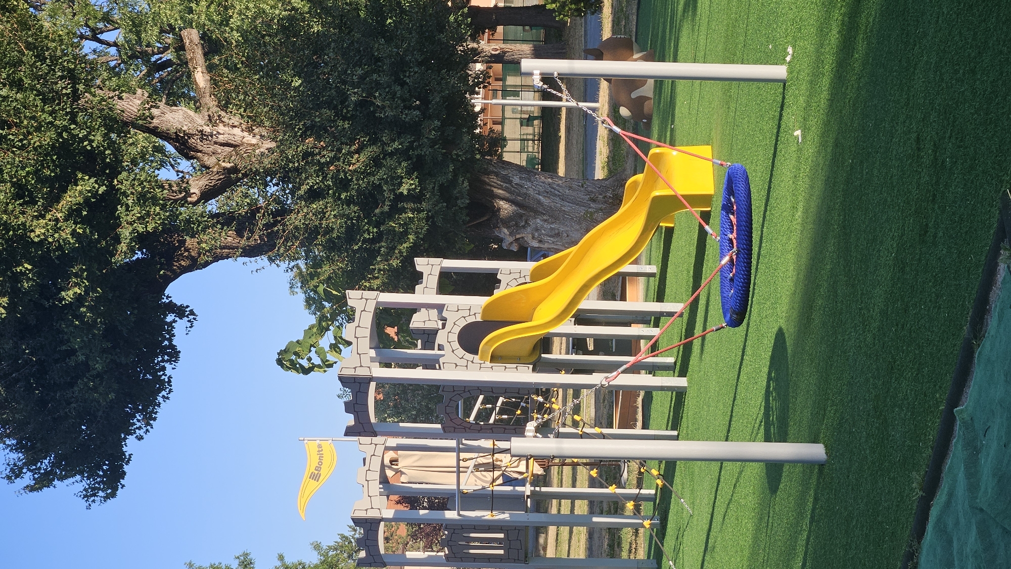 The image depicts an outdoor children's playground, specifically designed to resemble a castle, located in Prešov, Slovakia. The playground features a bright yellow slide, a blue nest swing suspended by red ropes, and a grey castle-themed climbing structure with turrets and battlements. A yellow flag with the text "Bonita" is attached to the castle structure. The ground is covered with artificial green turf, and several grey support poles are visible throughout the play area. In the background, a large, mature tree with dense foliage dominates the scene, with other trees and buildings suggesting an urban or park setting. A portion of a brown and white animal-shaped toy or decoration is visible to the right of the slide. The sky is clear and blue.