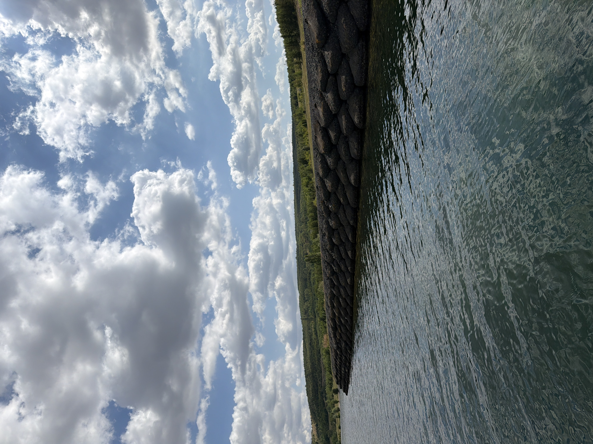 The image shows a body of water with ripples on its surface. Along the edge of the water is a man-made structure, possibly a breakwater or dam, constructed from large, dark, irregular stones stacked in layers. Beyond this structure, a green, tree-covered hillside rises gently. The sky above is a clear blue, dotted with numerous fluffy white and grey clouds. The overall scene depicts a natural landscape augmented by human engineering, under a dynamic sky. The location is identified as the water reservoir of Most, Czechia.