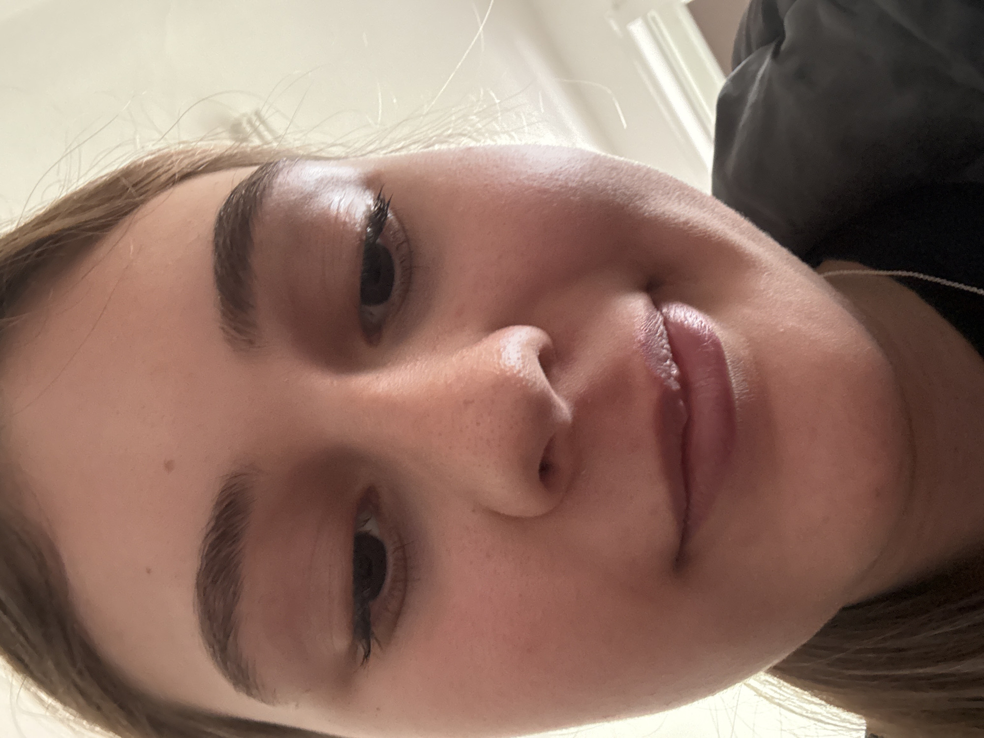 A close-up, low-angle shot of a fair-skinned young woman's face. Her gaze is directed downward towards the camera. She has brown hair visible along the edges of the frame. A black jacket or coat is seen on the right side of the image. The background includes a white wall. There is no indication of location (Panagyurishte, Bulgaria) in the provided image.
