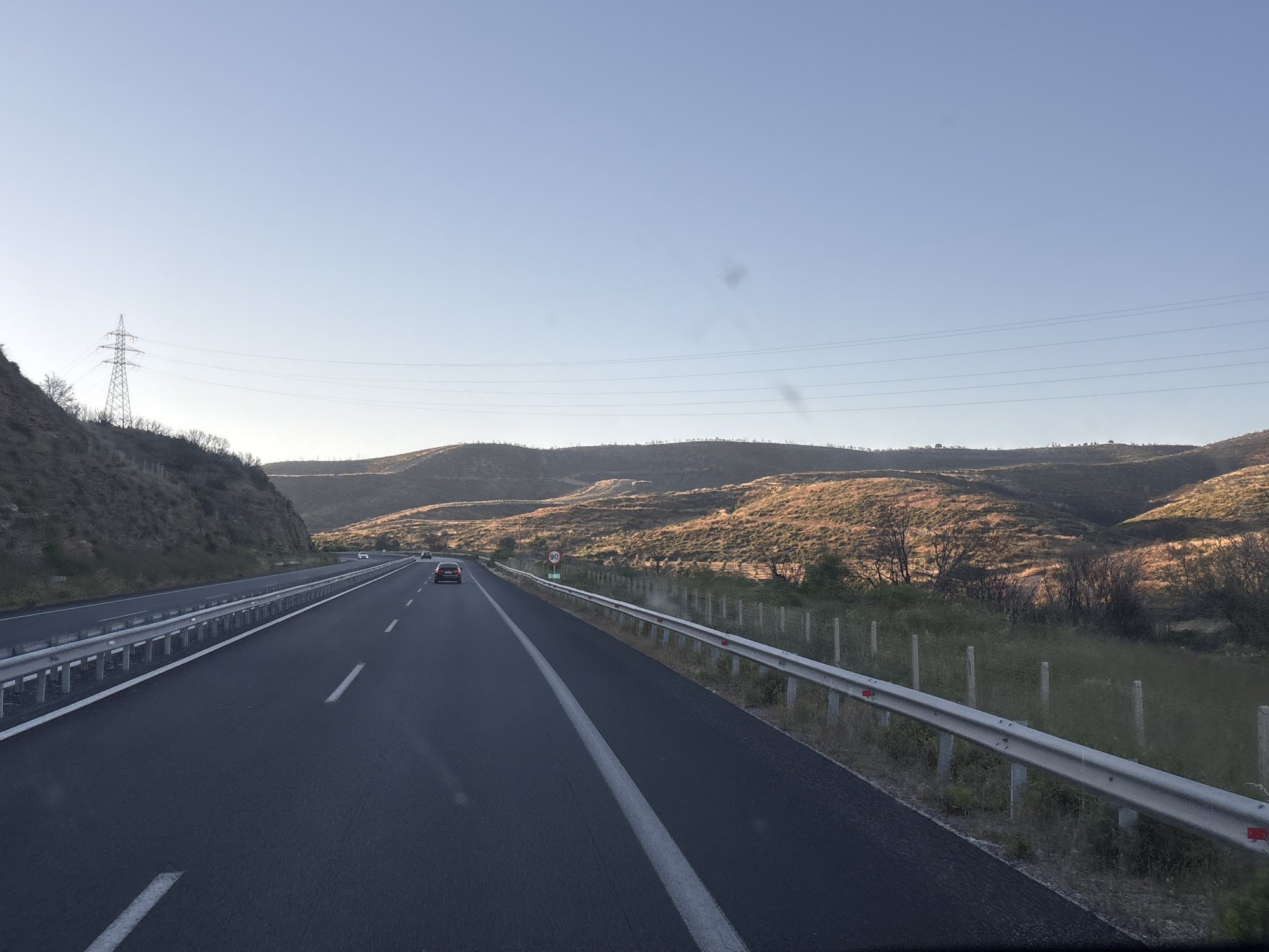 An outward-facing drone shot captures a highway with three lanes, two heading in the same direction, as it cuts through a valley in Avra, Greece. The road is dark asphalt, with a white line separating the lanes and a solid white line on the far left. A silver guardrail runs along the right side of the road. The landscape on both sides of the highway is a dry, hilly terrain covered with sparse, brown vegetation. In the distance, rolling hills are illuminated by the golden light of the setting sun, creating a warm glow. Power lines and a tall electrical tower are visible on the left side of the frame. There are no people visible, but two cars are driving on the highway. A circular speed limit sign with the number 90 is visible on the right side of the road.