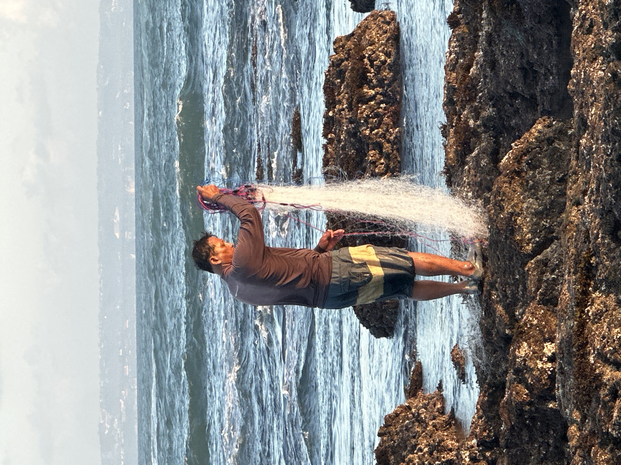 The image depicts a man standing on a rocky shore, engaged in fishing with a net. The man is wearing a long-sleeved shirt and shorts, and he is holding a fishing net that is spread out in the water. The sea is choppy with visible waves, and the sky is overcast. In the background, there is a hazy landscape of hills or mountains. The rocky shore is covered in barnacles and other marine life. The scene is likely located on the coast of North Kuta, Indonesia, given the visual characteristics of the shoreline and the ocean.