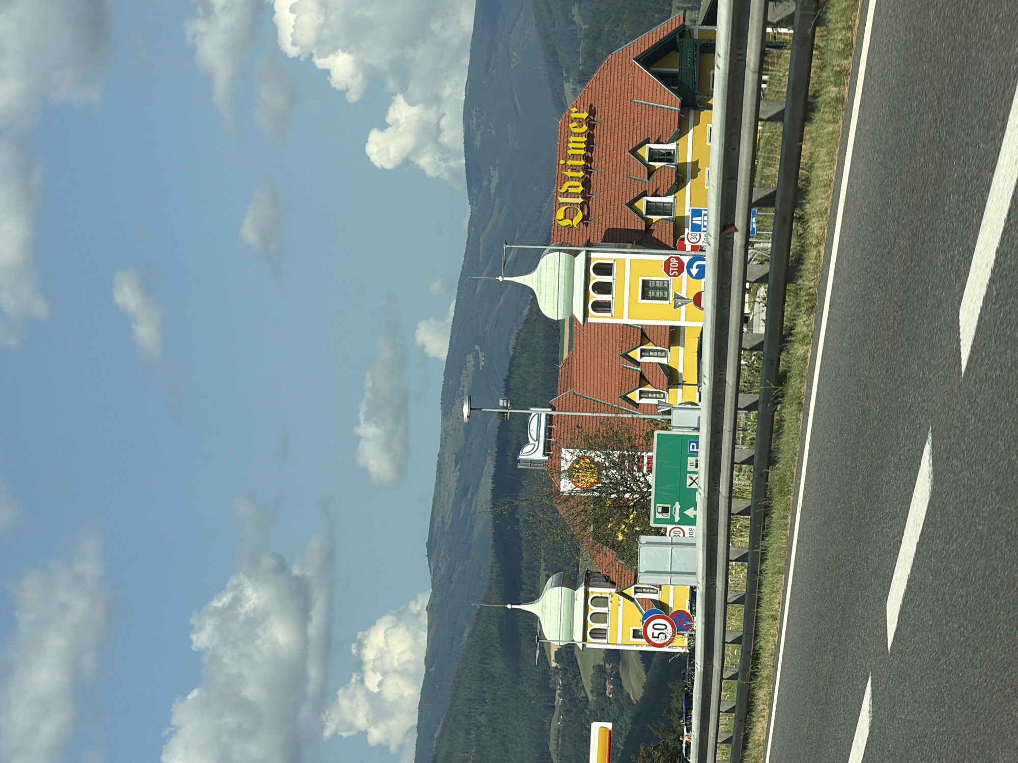 The image captures a roadside view of a building with a prominent "Oldtimer" sign in Maierhöfen, Austria. The building features a distinctive yellow facade with red-tiled roofs and green-domed towers. Various road signs are visible, including a speed limit sign of 50 and a STOP sign, indicating a nearby intersection or junction. A green sign with symbols for a gas station and parking is also present. The scene is set against a backdrop of rolling green hills and a partly cloudy blue sky. The foreground shows a section of asphalt road with white lane markings and a metal guardrail. There are no discernible people in the immediate vicinity of the building or on the road. The overall impression is that of a service area or rest stop along a highway.
