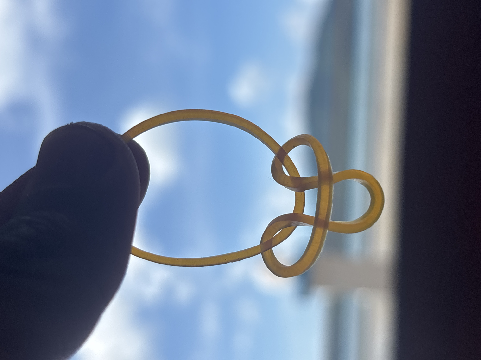 A close-up, silhouette shot captures a person's thumb and index finger holding a single, yellow rubber band. The rubber band is intricately twisted, forming a knot-like shape that resembles a stylized heart or infinity symbol. The background is a bright blue sky with scattered white clouds, softly blurred to emphasize the foreground subject. Below the sky, a hazy, out-of-focus line suggests a distant coastline or horizon, potentially the sea, with a dark, out-of-focus foreground strip implying a surface. The image appears to have been taken outdoors, possibly in Rawai, Thailand, given the clear sky and coastal hint.
