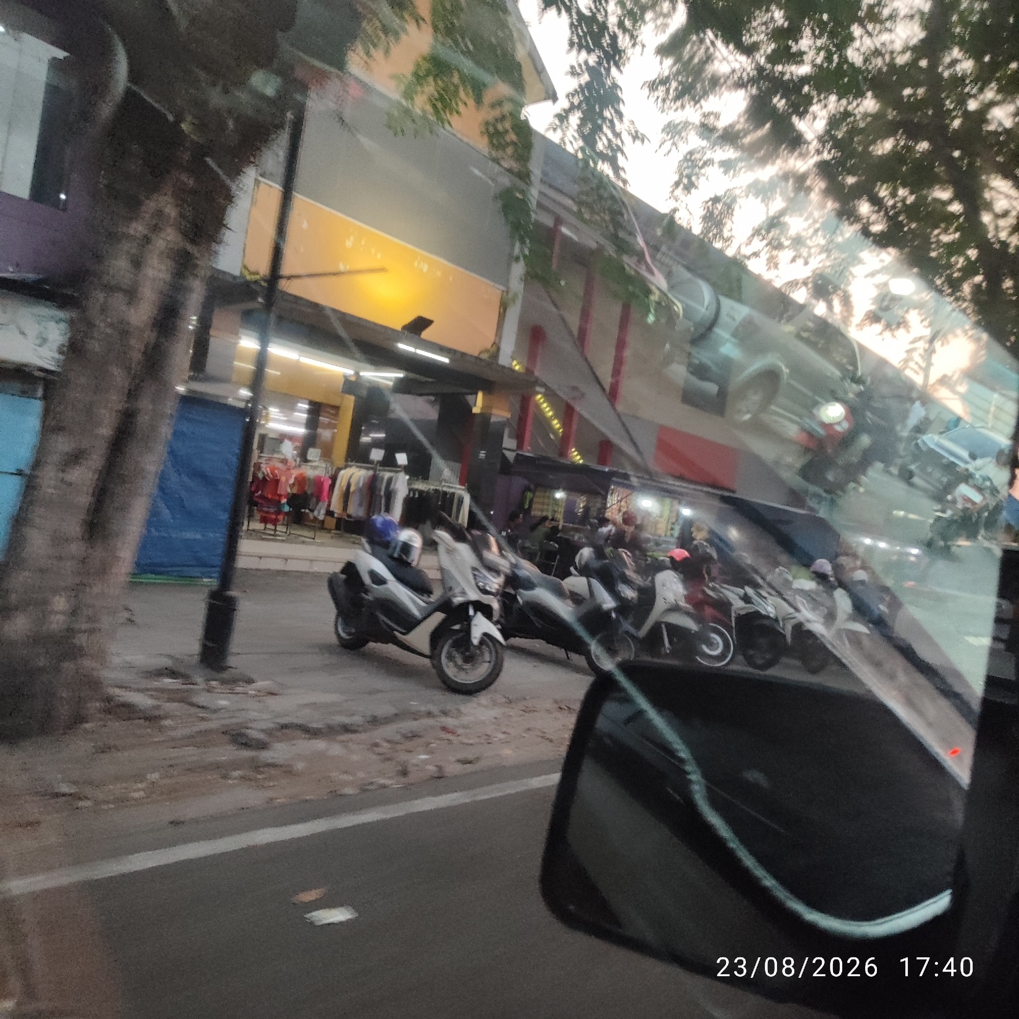This is a medium shot, taken from a vehicle, of a street scene in Tanjung Pandan, Indonesia. The foreground features a car's side mirror. Reflected in the mirror and the vehicle's windows are buildings, trees, and parked motorcycles. The buildings appear to be shops, one with a bright yellow facade and clothes displayed outside. Several motorcycles are parked on the side of the street. The timestamp at the bottom indicates August 23, 2026, at 17:40. The scene suggests a typical urban environment in Indonesia during the late afternoon.