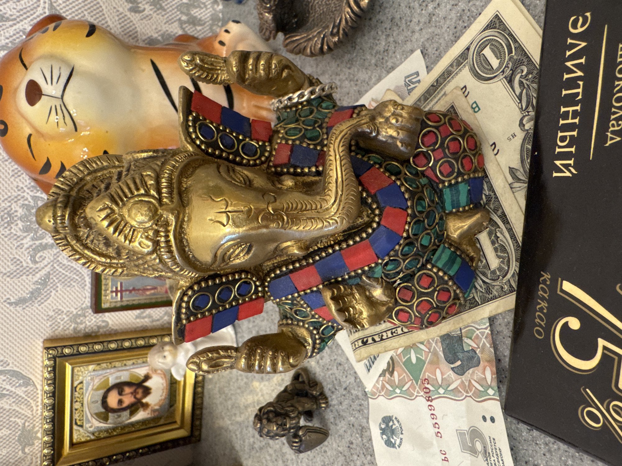 Here is a description of the image.
The image shows a variety of objects on a surface with a patterned gray tablecloth. Dominating the image is a golden statue of Ganesha, adorned with red, blue, and green stones. Next to Ganesha is a small ceramic tiger figurine. Also present are a framed icon of Jesus, a small metal figurine, U.S. dollar bills, Russian Ruble notes, and a box of dark chocolate with the words "75% cacao" on the front. The text "ЭЛИТНЫЙ ШОКОЛАД" in Russian is also visible on the box. The objects are arranged in a casual, somewhat cluttered manner. The location of the city of Unknown, Russia, is not mentioned or inferred from this image.