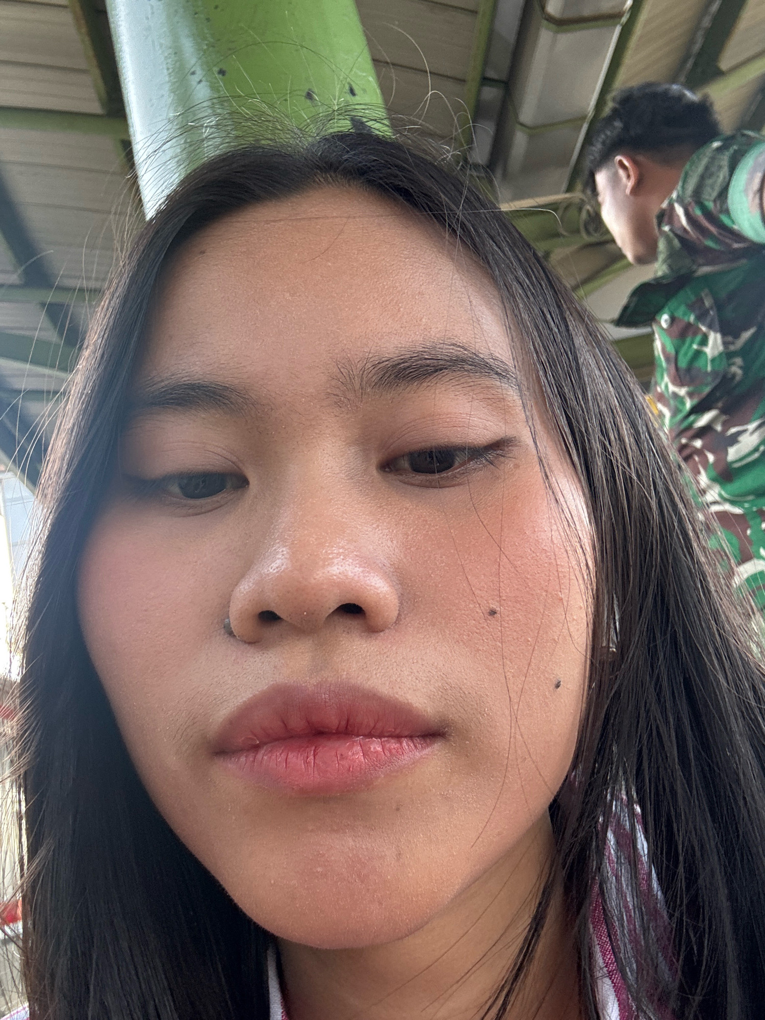 An extreme close-up selfie shows the face of a young woman from the chest up, angled slightly down and to her right. Her long, dark hair frames her face, with some strands falling across her forehead and cheek. Her skin appears smooth with visible pores and a few small moles. Her lips are slightly parted, and her eyes are looking down. She is wearing a red and white striped shirt, and a person in a green camouflage uniform is visible in the background, out of focus. The environment appears to be outdoors, with a green structure and what looks like corrugated metal roofing overhead. The lighting suggests it is daytime.