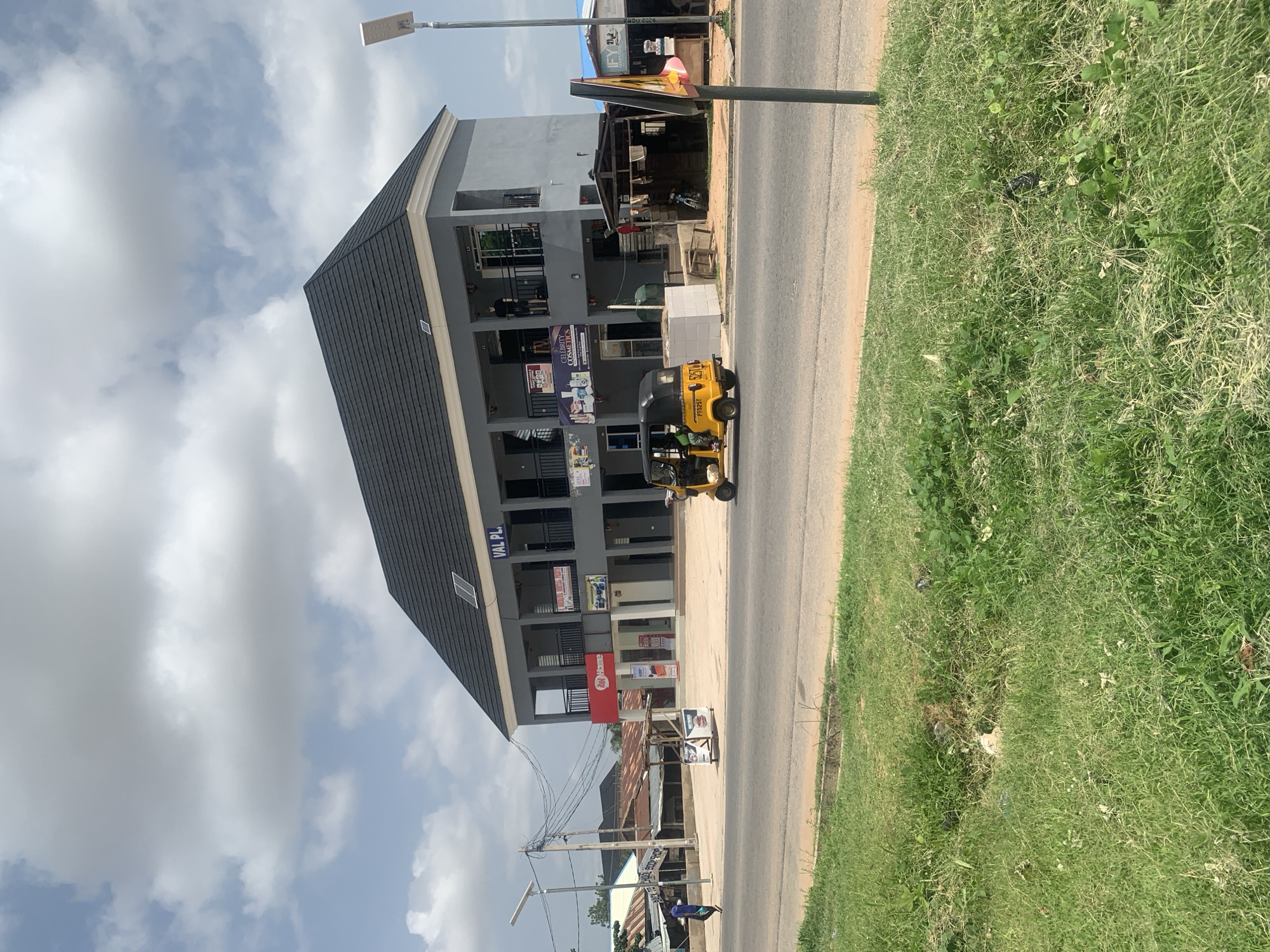 The image depicts a street scene in Jalingo, Nigeria, on a cloudy day. The foreground is dominated by a modern, multi-story building with a dark, sloped roof and grey exterior walls. The building has several levels with balconies and open windows. One sign visible on the building reads "VAL PL.". Below the main building, there are smaller shops with awnings and signage, one of which is red and says "RedHome". A yellow auto-rickshaw, also known as a "Keke Napep," is parked on the side of the road in front of the building. A person is visible inside the auto-rickshaw. Across the paved road, there is a patch of green grass with some scattered debris. In the background, another building with a corrugated iron roof is visible, along with telephone wires and a lamppost. The sky is filled with white, puffy clouds against a light blue background. The overall impression is a busy commercial area with a mix of modern and older structures.