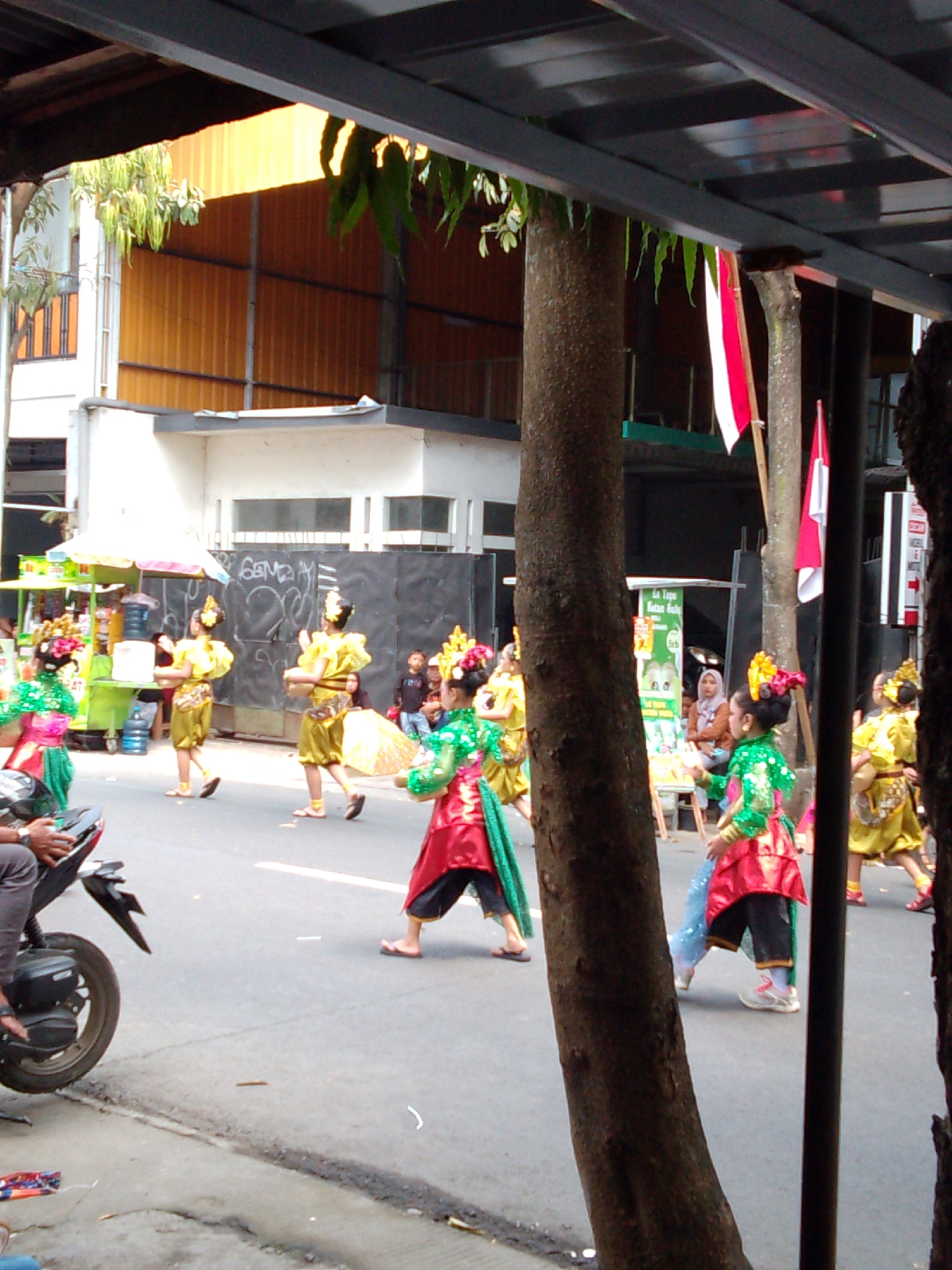 In Trenggalek, Indonesia, a group of children in traditional attire participates in a parade. The young dancers, adorned in vibrant yellow and green costumes with elaborate headdresses, move across a paved street. Some wear green shirts and colorful skirts, while others don yellow tops and skirts, their movements fluid and synchronized. A young girl in the foreground, wearing a sparkling green top and a red skirt, holds a blue sash. Behind them, a woman in a hijab watches the procession. The street is lined with buildings, including a white structure with dark-framed windows and an orange corrugated metal wall. Trees with textured bark stand on the roadside, partially obscuring the view. A motorbike is parked on the left side of the street. Indonesian flags, red and white, are displayed on poles, adding to the festive atmosphere. The scene captures a cultural event unfolding in the city.