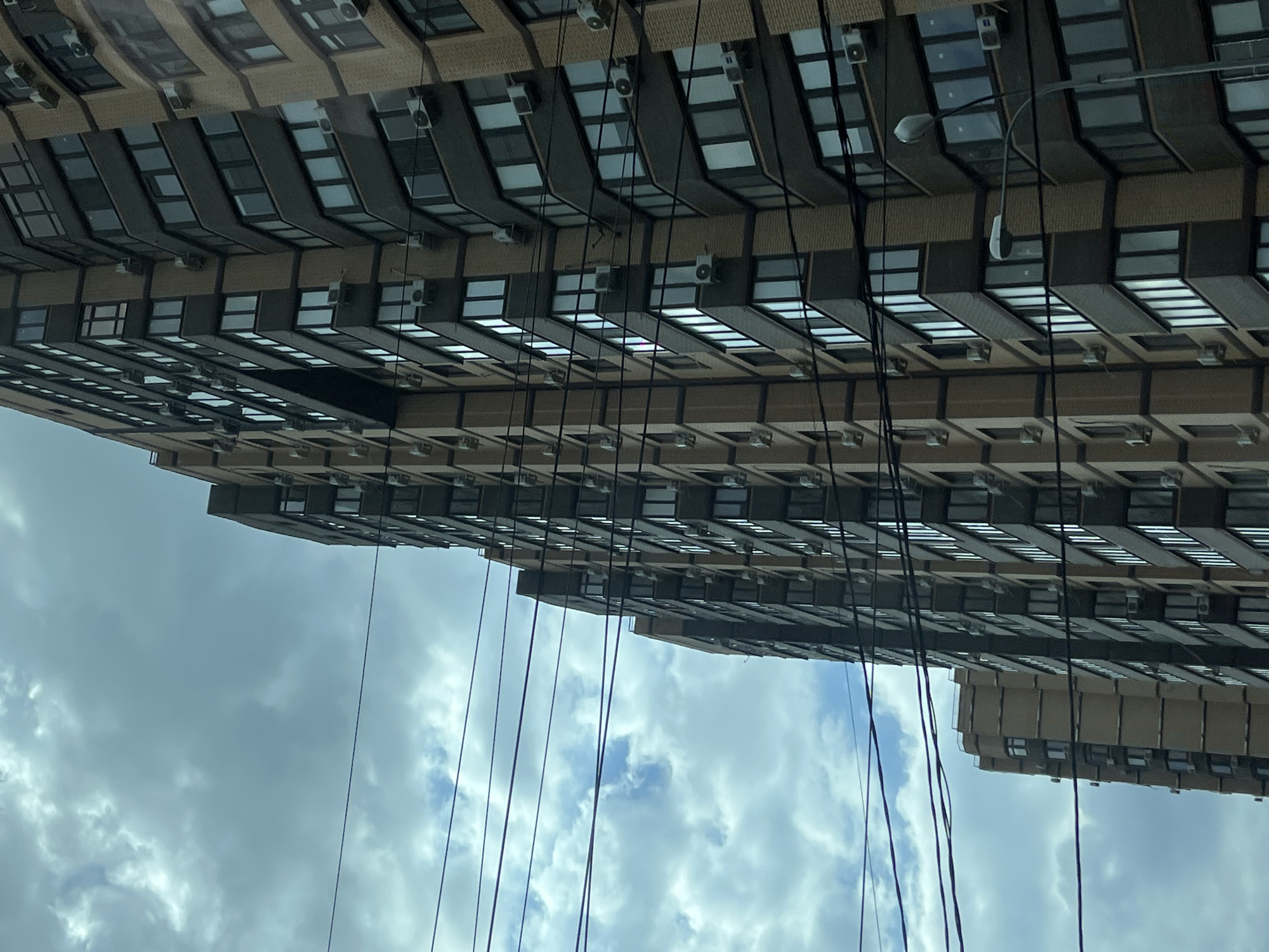 This media file presents an urban scene dominated by a multi-story building, oriented with a 90-degree clockwise rotation. The building occupies the top and right portions of the frame, appearing to stretch diagonally across. Its facade features repetitive patterns of windows and exterior panels in shades of light brown brick and dark grey. Numerous white outdoor air conditioning units are mounted beneath almost every window, indicating individual climate control for many dwelling units or offices.
A dense array of thin, dark wires hangs vertically across the image, obscuring parts of the building and the cloudy sky. A single grey street lamp, with an extended arm, is visible in the upper right, attached to the building structure. The bottom and left sections of the image are filled with a bright, cloudy sky, featuring scattered white and grey clouds.
The scene depicts a static architectural view in an urban environment, likely during the daytime given the bright sky. No specific activity or event is taking place. There is no legible text visible anywhere in the image. The location context provided is Krasnodar, Russia.