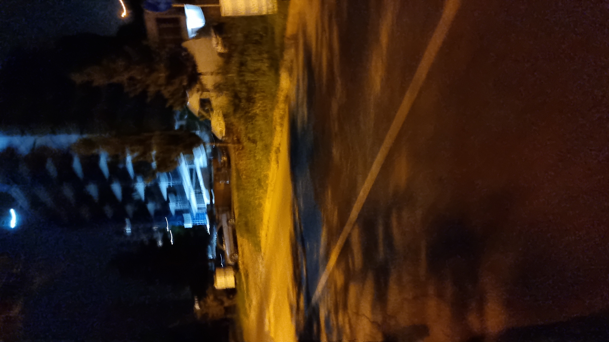 An out-of-focus, nighttime, street-level view of a building and a road in Tbilisi, Georgia. The building appears to be a multi-story structure with illuminated windows, possibly under construction or renovation given the scattered lighting and general blurriness. The road in the foreground is asphalt with a white line marking. There are trees and some parked vehicles visible on the side of the road. The overall impression is of a blurry, indistinct urban scene at night.