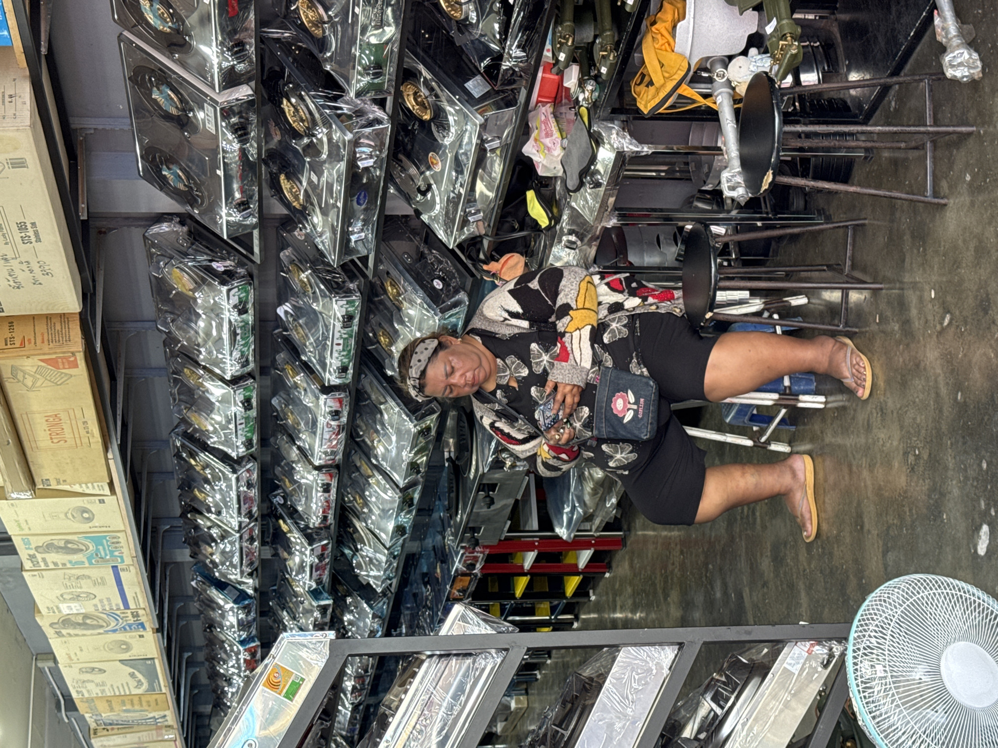 In Ranong, Thailand, a woman sits on a black stool in what appears to be a retail store or warehouse. She is wearing a black headband, a patterned long-sleeved shirt with black, white, and red colors, and black shorts. She holds a smartphone in her hands and has a small denim purse with a pink flower emblem slung over her shoulder. The store is filled with rows of new gas stoves, each packaged in clear plastic. The shelves are stocked high with these appliances. In the foreground, a white fan is visible on the floor. The environment suggests a commercial setting with a focus on kitchen appliances.