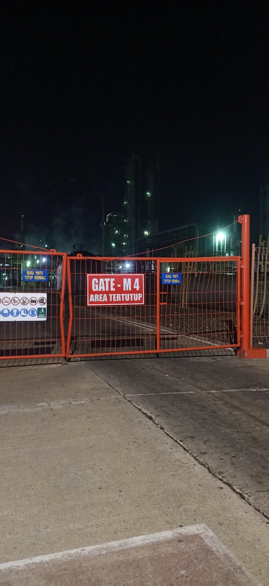 The image shows an industrial facility at night, possibly in Palohlada, Indonesia, based on the Indonesian text on signs. An orange metal fence with a wire mesh infill blocks a paved pathway. In the center of the fence, a red sign reads "GATE - M 4 AREA TERTUTUP" (GATE - M 4 AREA CLOSED). To the left and right of this central sign, blue signs are attached to the fence. The left blue sign is partially visible and reads "BUKA PINTU TUTUP KEMBALI" (OPEN GATE CLOSE AGAIN). A white sign with several circular icons depicting safety regulations is attached to the left side of the fence. In the background, large industrial structures with illuminated lights are visible against a dark sky. There are no people or vehicles visible in the image. The ground in the foreground is concrete with visible cracks.