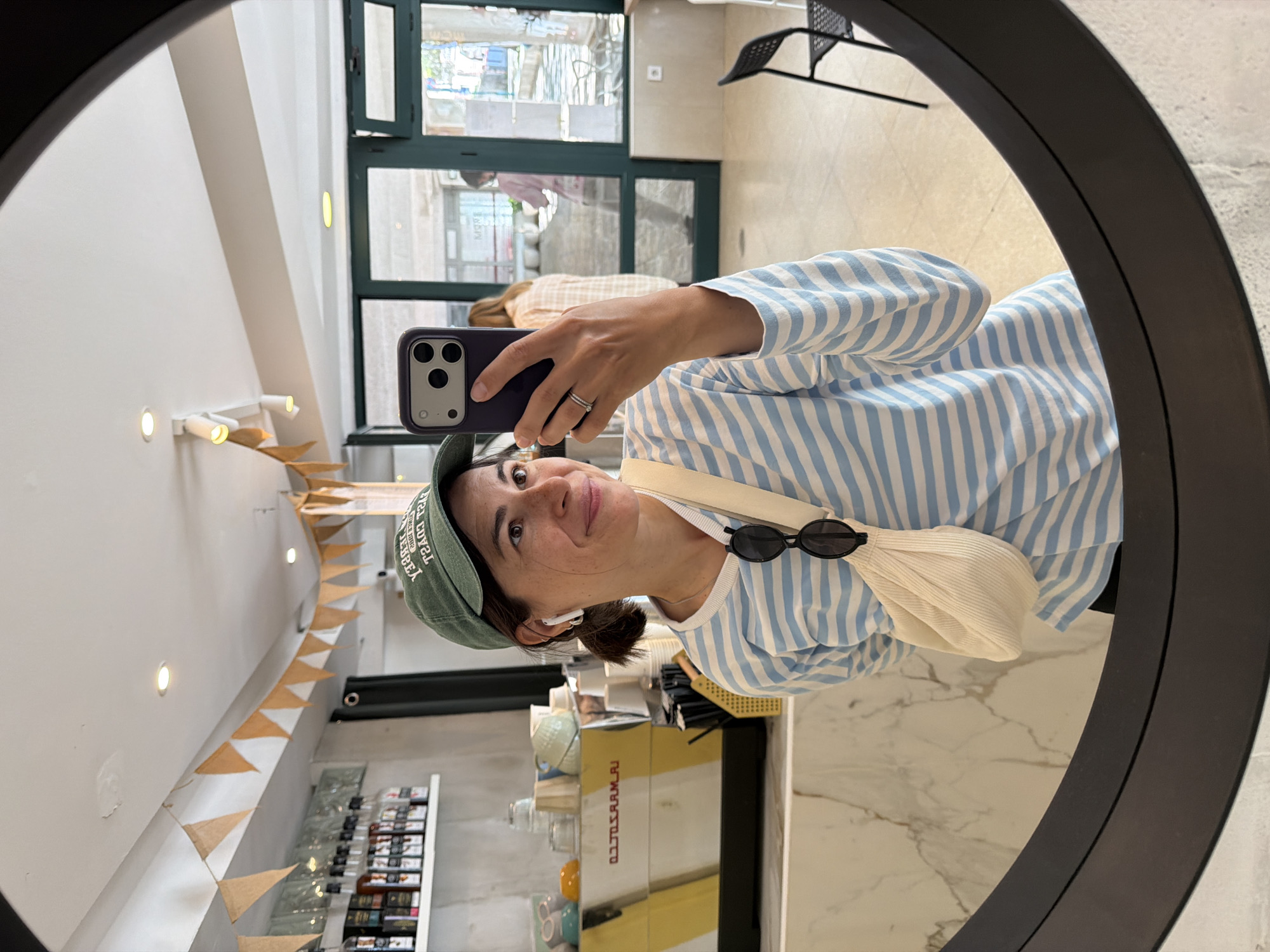A woman is taking a selfie in a round mirror. She is wearing a blue and white striped shirt, a green baseball cap with white lettering, and an off-white crossbody bag. She is holding a smartphone in her right hand. In the background, there is a shop interior with retail products on shelves, a glass door, and a chair. The shop is located in Budva, Montenegro.