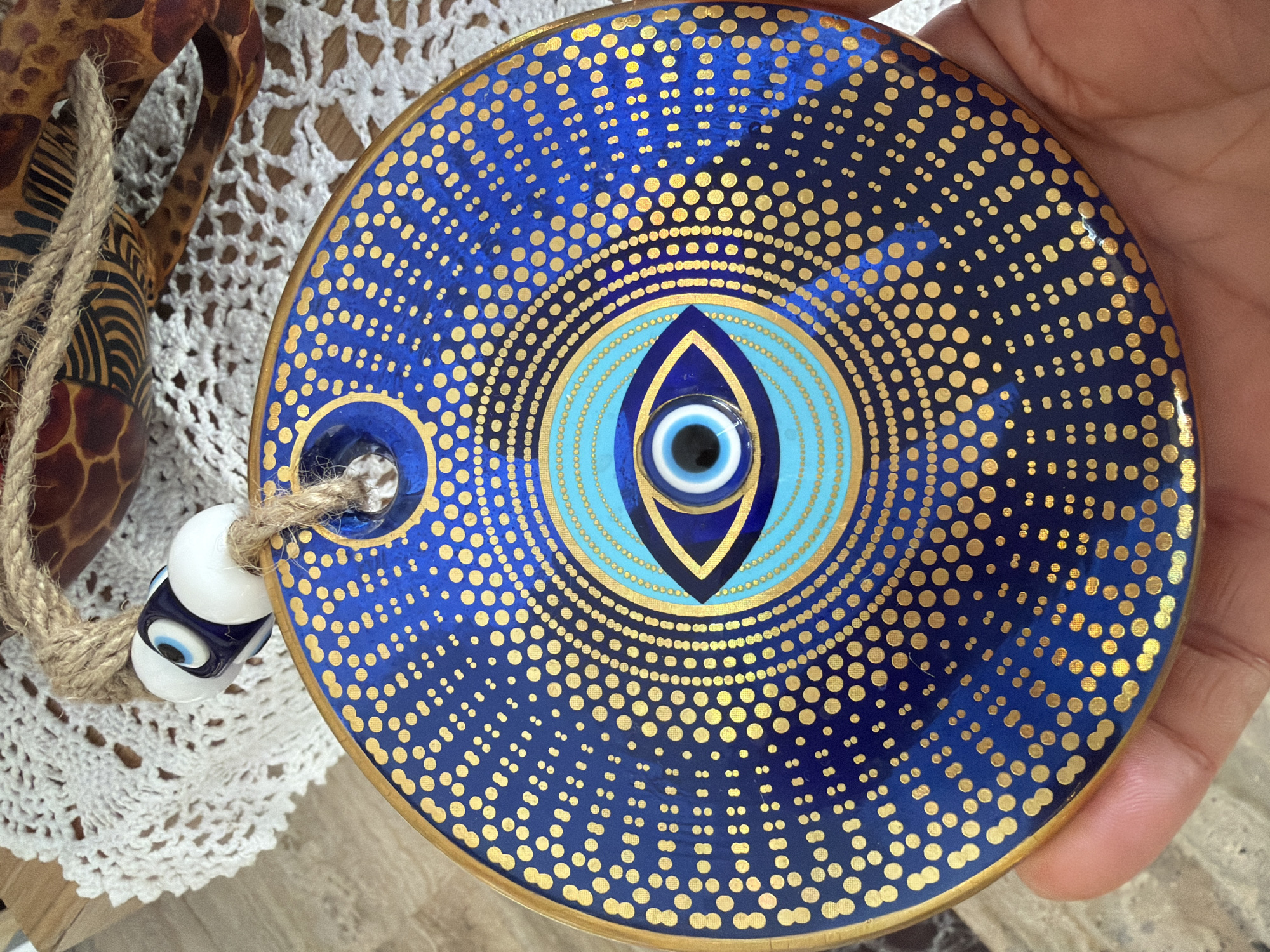 A hand holds a round, deep blue ceramic evil eye ornament adorned with gold dots and concentric circles. In the center of the ornament is a stylized eye design with a blue iris and a black pupil, surrounded by lighter blue rings. The ornament is suspended from a rough jute rope, from which two beads hang: one white with a blue and black eye design, and another solid white bead. The ornament is placed on a white, crocheted doily, behind which a portion of a wooden table is visible. In the background, a carved wooden object with a giraffe-like pattern is partially visible. The scene appears to be indoors, possibly a souvenir shop or home, given the decorative nature of the objects and the presence of a doily. The location is stated to be Marmaris, Turkey.