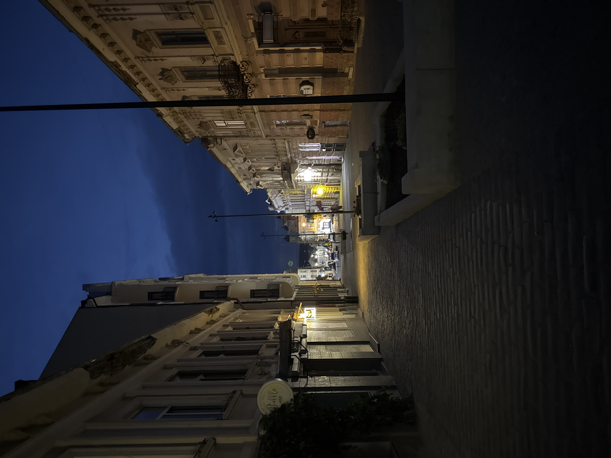The image depicts a cobblestone street in Chernivtsi, Ukraine, at twilight. On the left, a building features a sign with "Bubble me" and is adorned with ivy. The building on the right showcases ornate architectural details and balconies. The street is illuminated by warm streetlights, casting long shadows on the ground. In the distance, a few cars are visible, along with more buildings and streetlights. The sky is a deep blue, with faint clouds. The scene appears quiet and tranquil.
