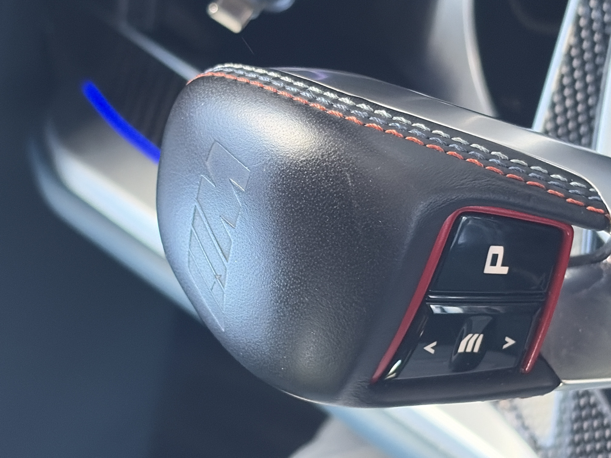 The image shows a close-up of a car's gear shifter. The shifter is black with a leather-like texture and features an embossed logo on its side. The top of the shifter is stitched with orange and gray thread. A red-trimmed panel on the shifter houses two buttons. The top button features a symbol that appears to be the letter "P," possibly indicating a parking or electronic parking brake function. The lower button has symbols indicating directional controls, with arrows pointing left and right, flanking a symbol consisting of three vertical lines. The background is blurred, revealing parts of the car's interior, including a blue light strip and a carbon fiber-like panel on the steering wheel. The image does not contain sufficient information to determine the location in Shumen, Bulgaria.