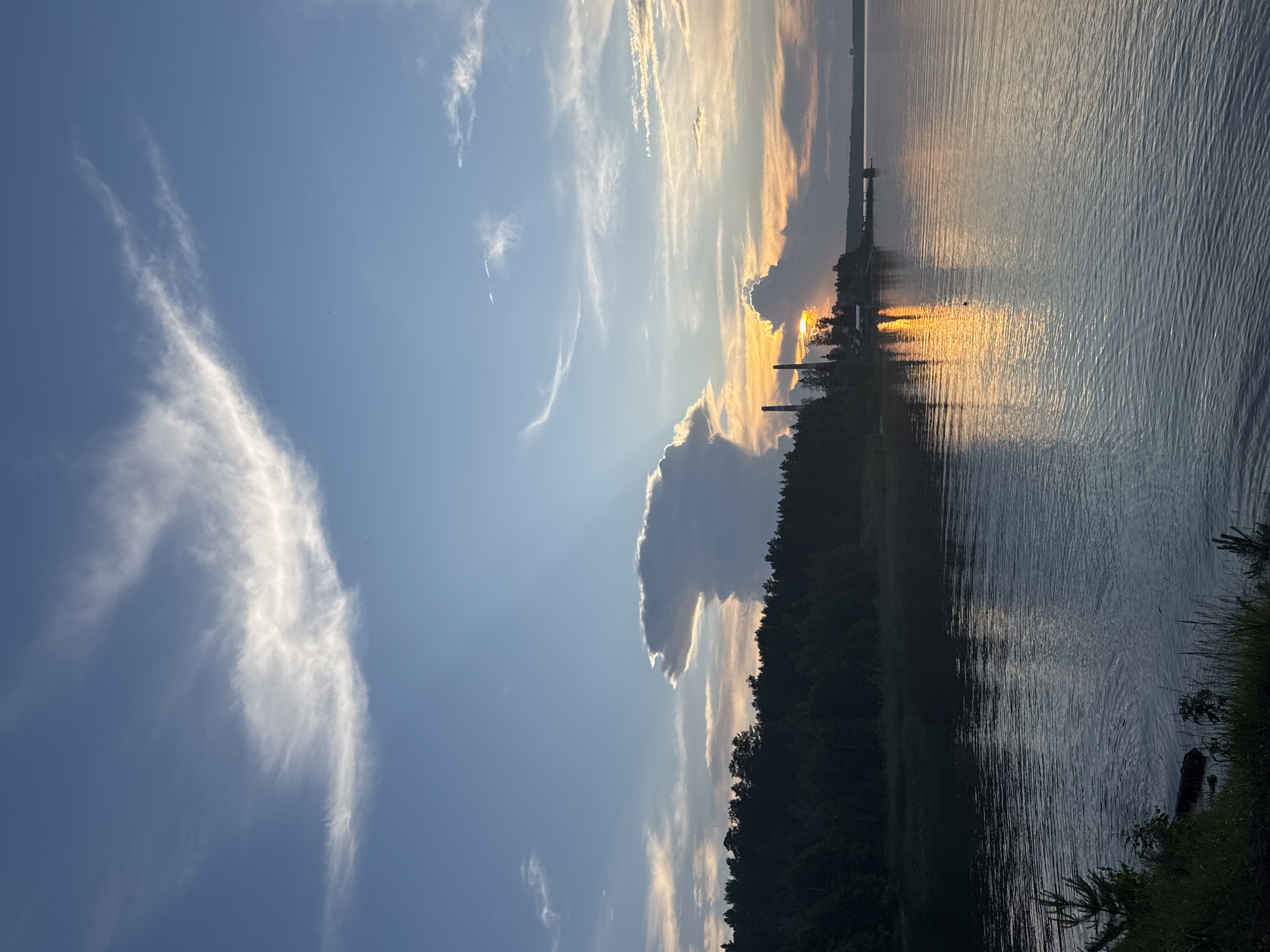 This image depicts a sunset over the Kama River in Chaikovsky, Russia. The sky is a brilliant canvas of blues and oranges, with wispy white clouds stretching across the horizon. Sunlight streams through a gap in the clouds, casting a golden glow on the water. On the left side of the frame, a dense line of dark green trees forms the shoreline. In the distance, silhouetted against the vibrant sky, are several industrial structures, including tall, slender smokestacks. The calm water of the river reflects the colors of the sunset, creating a mirror image of the sky. The foreground shows the grassy bank of the river with a few small pine trees. The scene is tranquil and serene, capturing the beauty of a natural landscape juxtaposed with industrial elements.