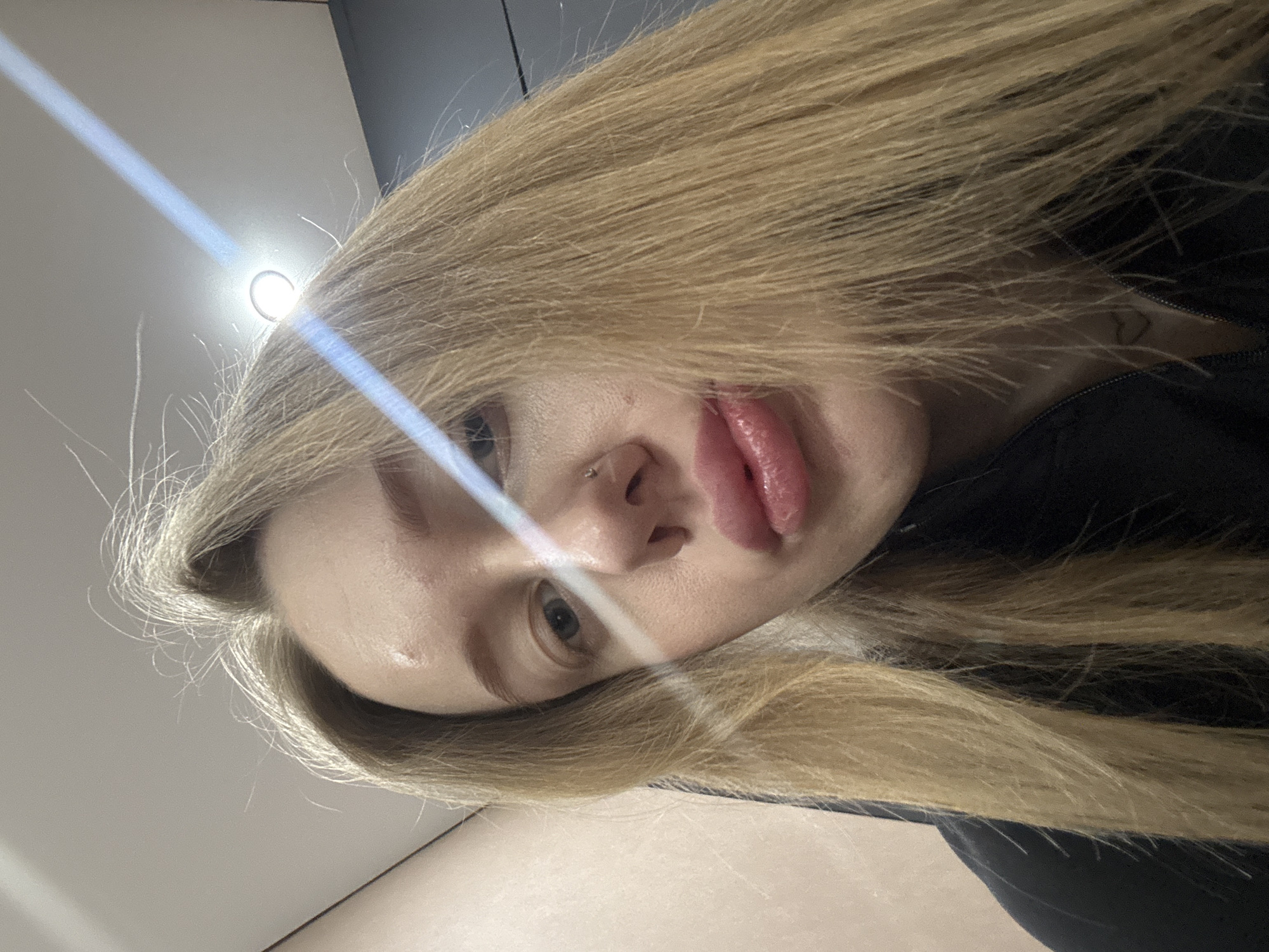 A close-up, angled shot of a person's face with long blonde hair partially obscuring the view. The individual has fair skin, a nose piercing, and pursed lips. A bright light source is visible in the upper left corner, casting a strong streak of light across the face. The background appears to be a simple interior with light-colored walls. There is no evidence to suggest the location is in Azov, Russia.