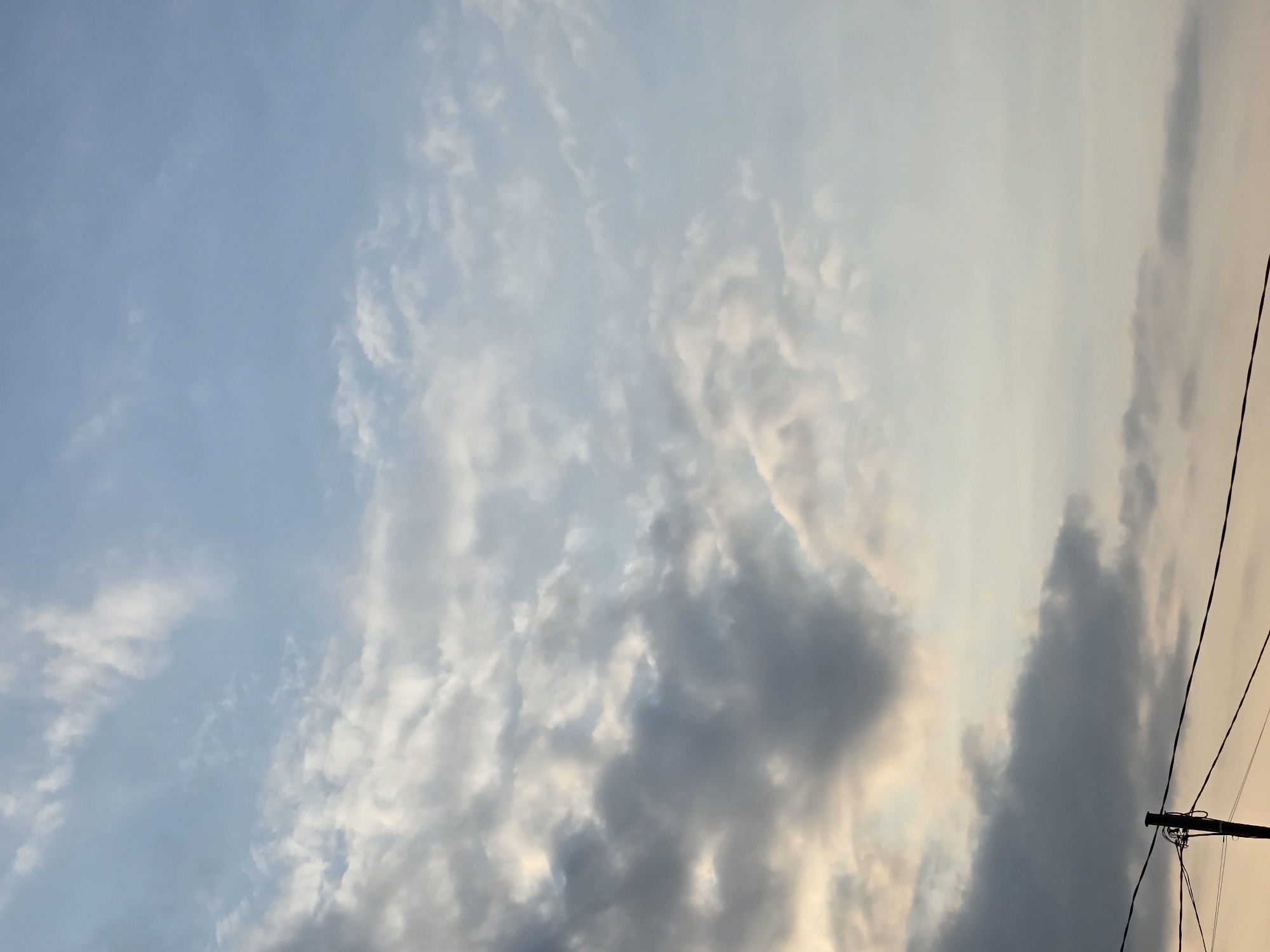 The image shows a sky with scattered clouds at different altitudes, some darker and denser, others wispy and light. The sky is a gradient of blue and pale yellow, suggesting either dawn or dusk. In the lower part of the frame, a black wooden pole is visible, supporting several electrical wires that stretch horizontally across the image. These wires are thin and dark against the brighter sky. The scene appears to be outdoors, likely in a semi-urban or rural setting due to the presence of utility poles and wires. The overall atmosphere is calm and atmospheric.