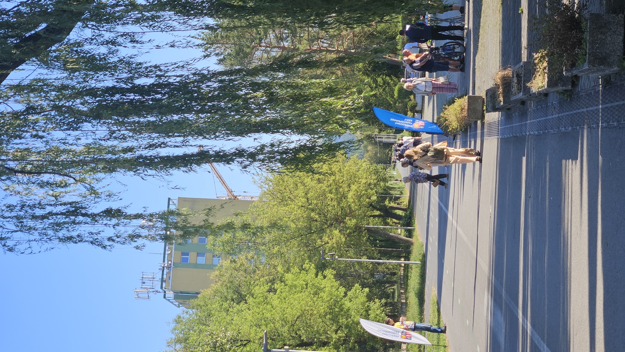 The image depicts a street scene in Prešov, Slovakia, likely in an urban park or along a designated pathway. The foreground features an asphalt pathway with white lines, indicating a lane for walking or cycling. Sunlight casts long shadows from trees onto the path. On the right side of the path, there are concrete planters with sparse vegetation.
In the mid-ground, several people are walking or standing. A woman in light brown trousers and a backpack is walking away from the camera. To her left, a group of people are gathered, some standing and others appearing to converse. A blue flag with text is positioned behind this group. Further down the path, more individuals are visible, suggesting a public event or gathering.
The background reveals a green building with multiple floors and windows, topped with cellular antennas. A construction crane is also visible in the distance. Lush green trees line the left side of the pathway, with one particularly large weeping willow tree partially obscuring the view with its long, hanging branches. The sky is clear and blue. A white flag with text is positioned on the far left of the pathway.