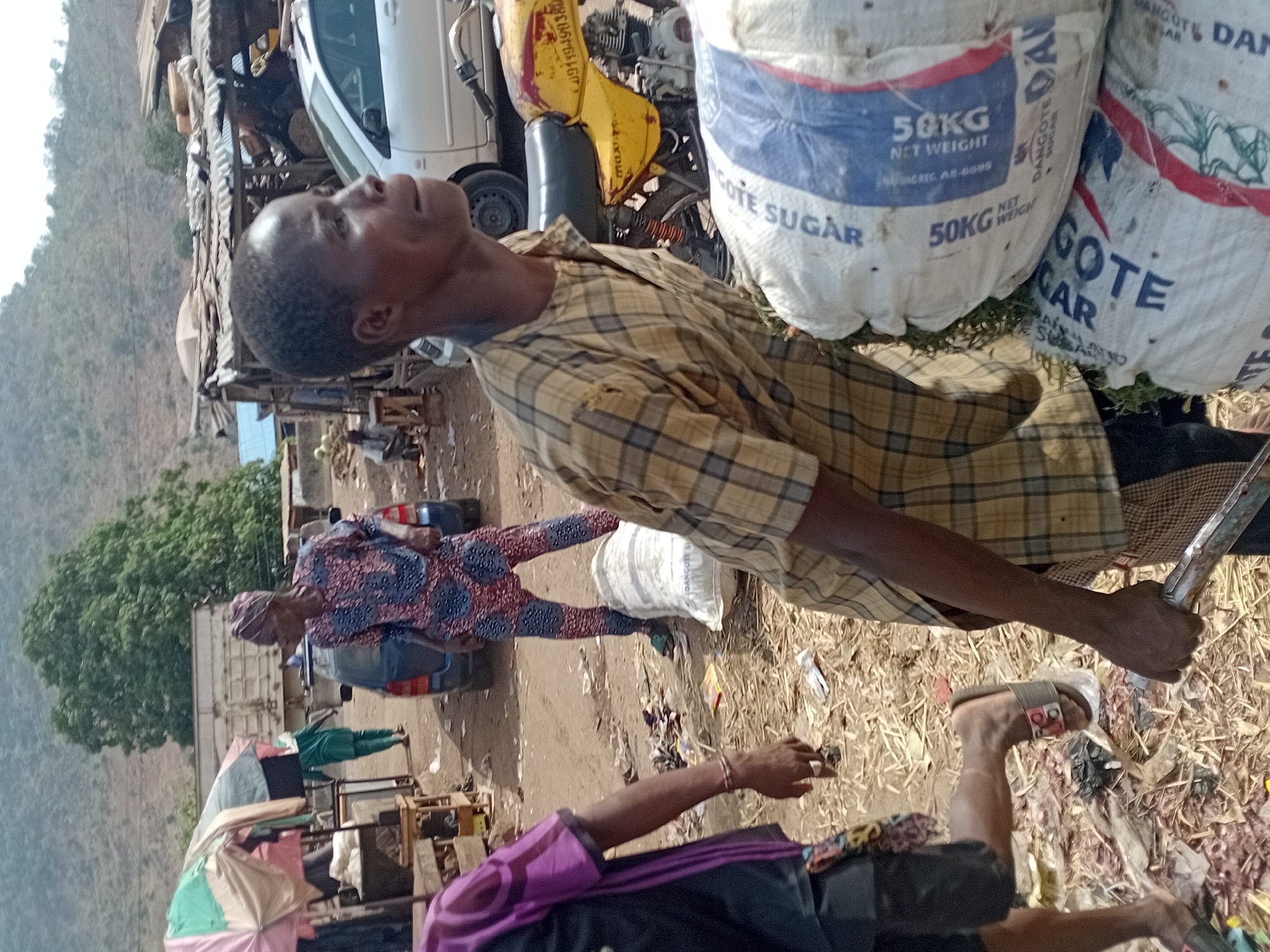 The image depicts an outdoor scene in Lokoja, Nigeria, exhibiting signs of commercial activity and potential junkyard elements. A man in a plaid shirt stands in the foreground, looking upwards. To his right are several large sacks of Dangote Sugar, each marked as 50kg net weight. In the background, a white vehicle is visible amongst what appears to be scrap metal and engine parts. Other individuals are present, one wearing a colorful patterned outfit. There are makeshift structures, possibly market stalls, under umbrellas. The ground is littered with debris and straw. In the far background is a mountain range.