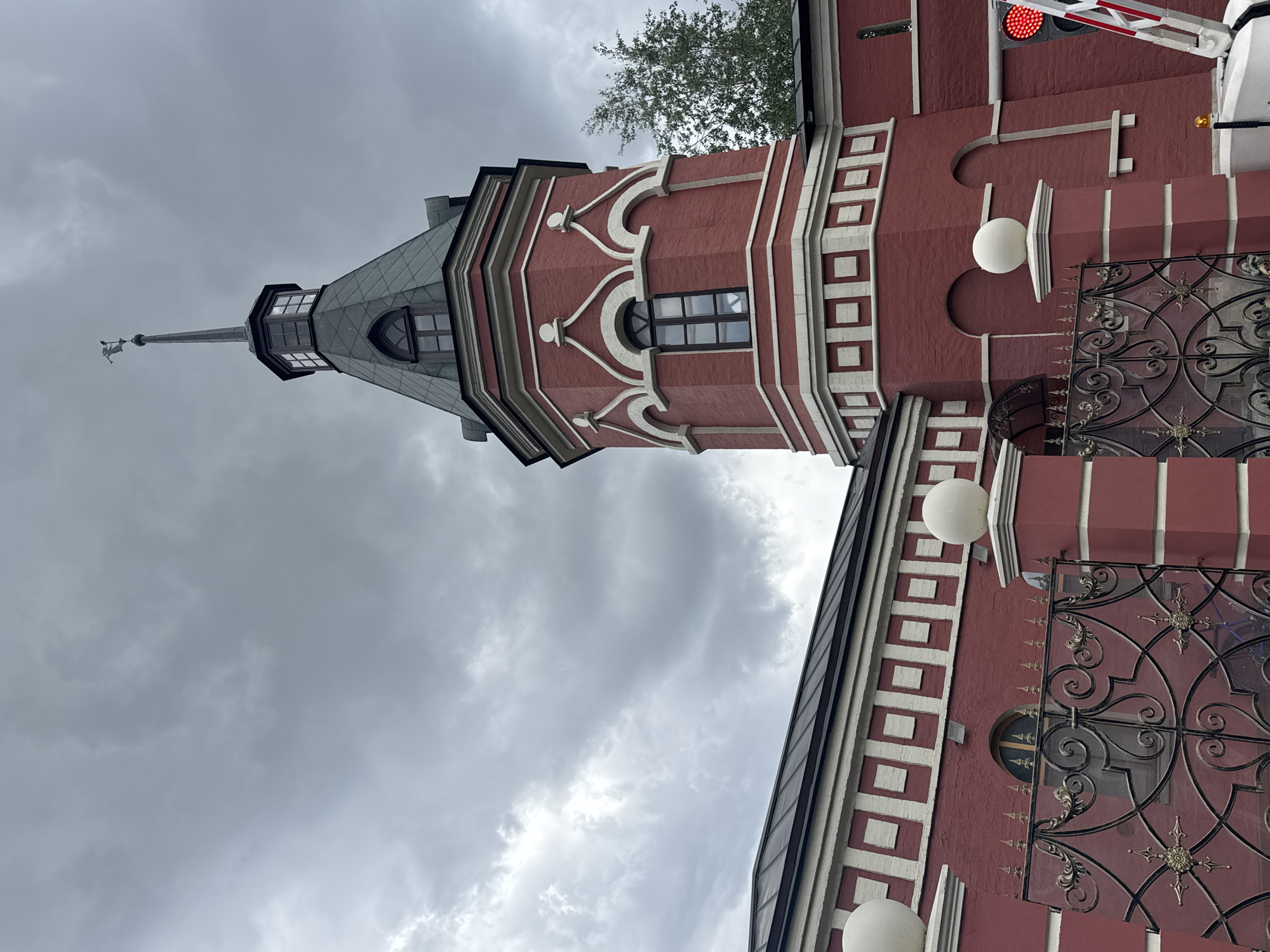 The image shows a fragment of the Izmailovo Kremlin in Moscow, Russia. The architectural style is an imitation of Russian architecture from the 16th-17th centuries. The structure visible in the picture is predominantly red brick with white decorative elements. A tower with a conical roof rises prominently. There is ornate metalwork on the building. Above the building, the sky is overcast with gray clouds. The building also includes features such as windows, spheres, and a weather vane. There's a red traffic light visible at the top right.