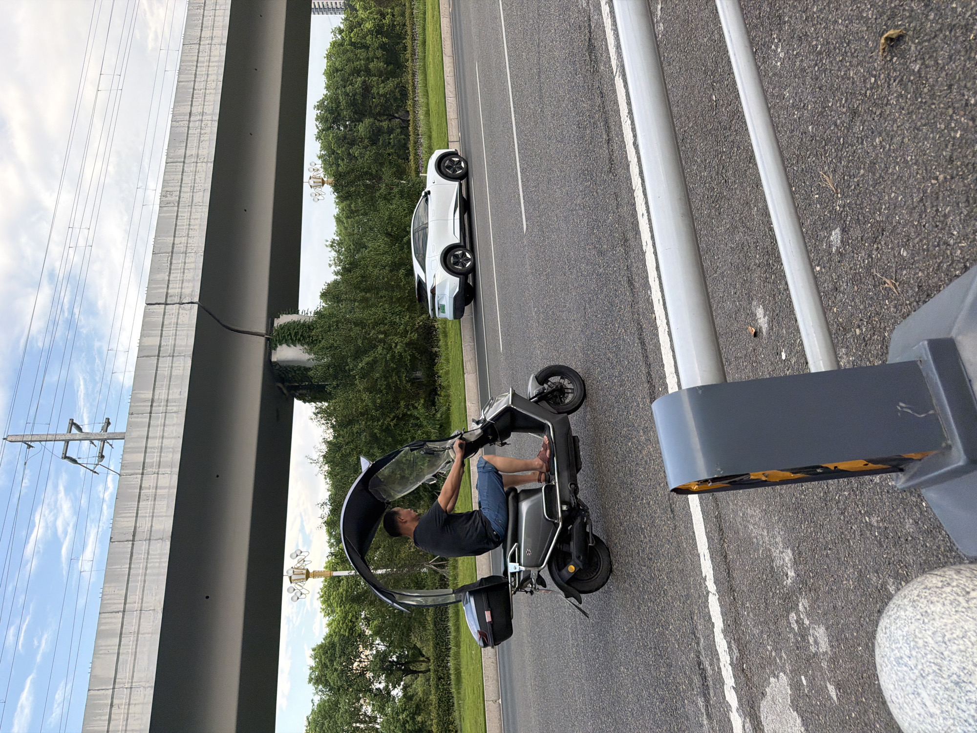 In Jiangjin, China, a man rides a gray electric scooter with a canopy and windshield on a paved road. He is wearing a black t-shirt and blue shorts. To his right, a white electric car is parked on the side of the road. Overhead, a concrete overpass is visible with power lines running across the sky. The background consists of green trees and a cloudy sky. In the foreground, a grey traffic barrier with yellow reflective tape is partially visible.