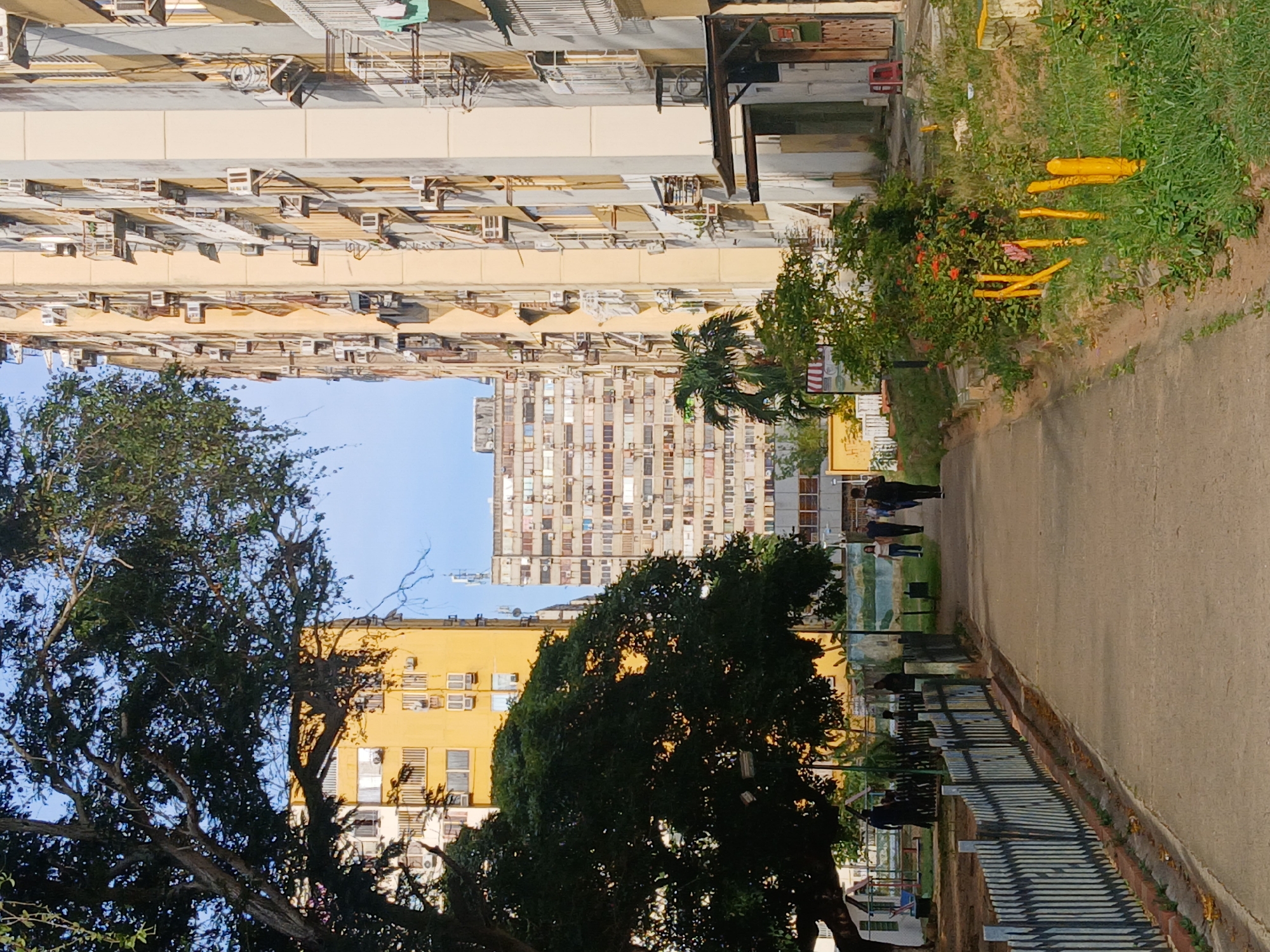 The image depicts a street scene in a densely populated area with tall buildings. The buildings are primarily light beige or yellow and have numerous air conditioning units attached to their facades. A paved pathway runs through the center of the image, lined with trees and vegetation. People are walking along the path. A low fence runs along the right side of the path, separating it from a grassy area. The sky is visible in patches between the buildings and trees, appearing light blue. Yellow safety poles can be seen in the grassy area. It is not possible to confirm the location in the city of Ciudad Guayana, Venezuela from the given image.