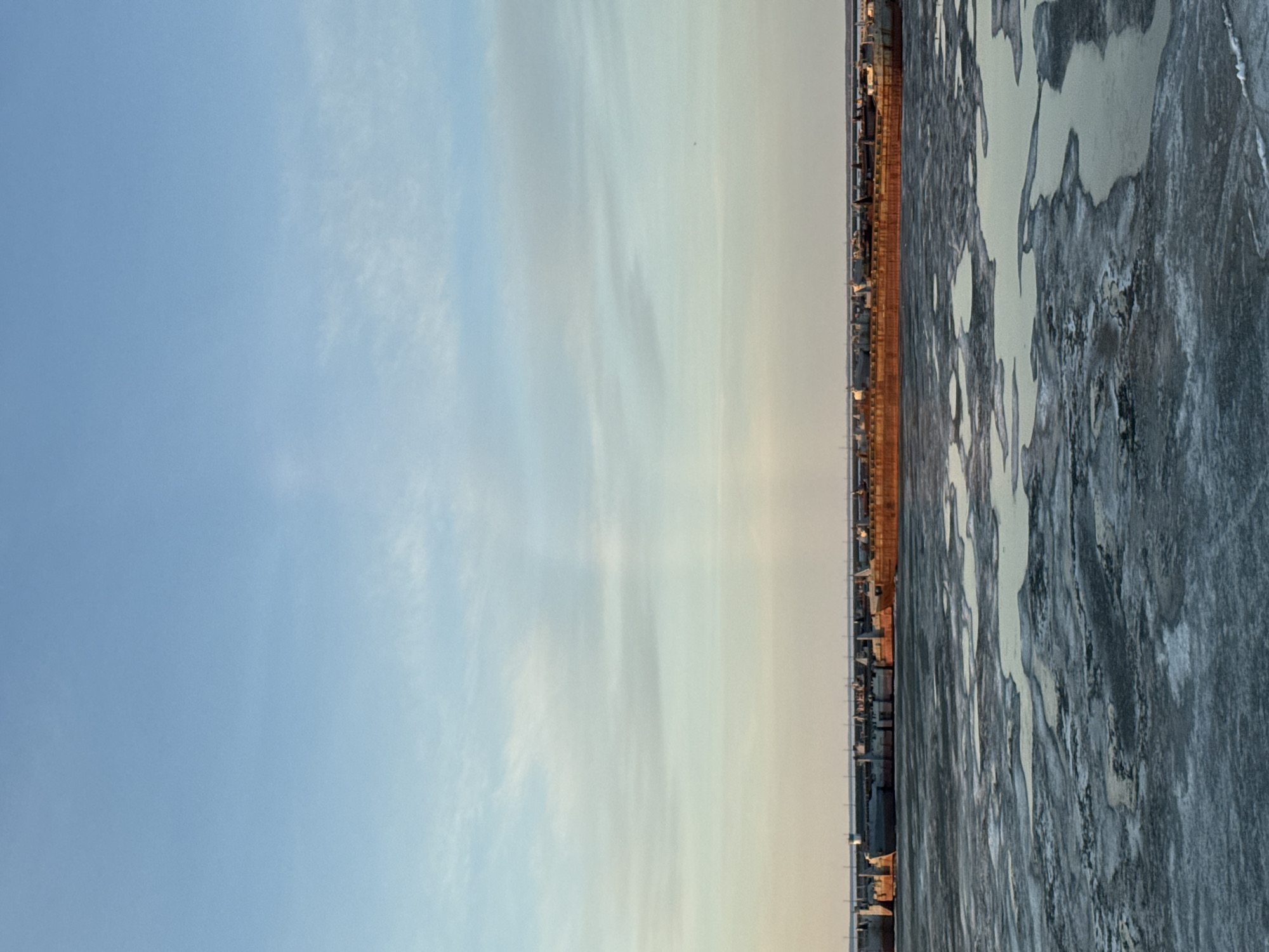 This landscape image depicts a vast winter scene under a clear sky. The upper two-thirds of the frame are filled with the sky, which transitions from a deeper blue at the top to a lighter, warmer pale blue or yellowish tone near the horizon. Sparse, wispy white clouds are visible, adding subtle texture to the expansive blue.
The lower third of the image is occupied by a wide, partially frozen body of water. The surface is characterized by a mosaic of light-colored, irregularly shaped ice patches contrasting with darker areas, which may be open water or thinner ice. The texture of the ice appears rough and fragmented.
A thin, dark line along the horizon separates the water from a distant landmass, where faint structures, possibly buildings or industrial elements, are discernible. These structures are subtly illuminated by a soft, warm light, suggesting either sunrise or sunset. The presence of significant ice confirms cold, winter conditions.
The scene conveys a sense of peaceful vastness and natural solitude. No individuals, prominent foreground objects, or visible text are present. Considering the location context, this is likely a view of the Kama River or Votkinsk Reservoir near Chaikovsky, Russia, during the colder months.