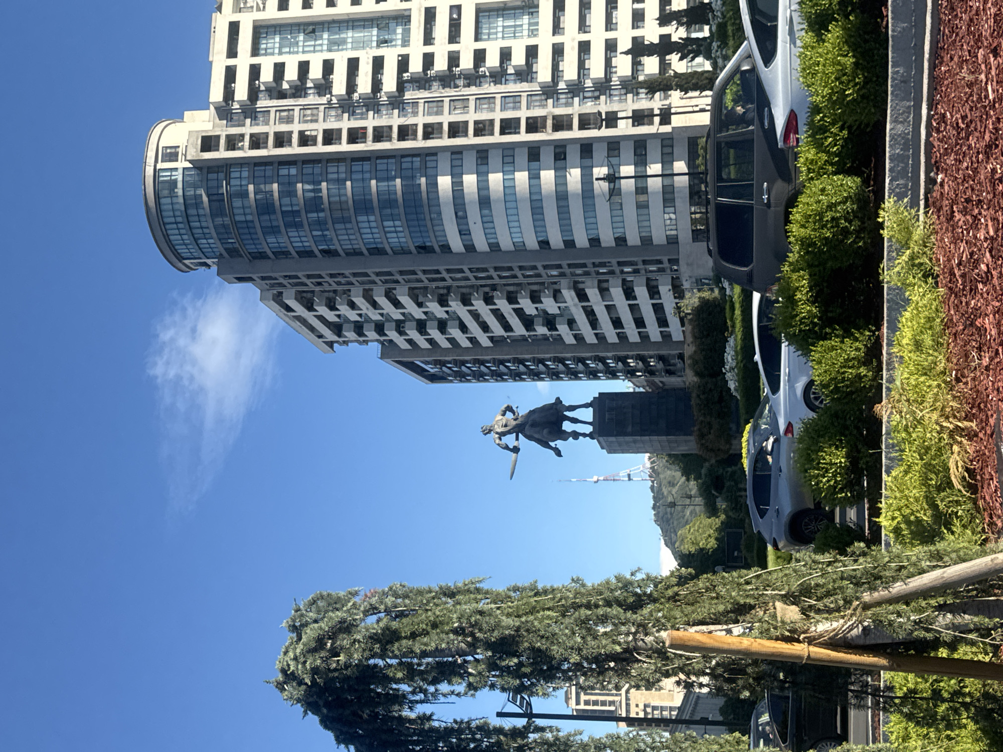 In Tbilisi, Georgia, on a clear day with a few wispy clouds, a statue of a warrior on horseback is prominently displayed in the foreground, holding a sword. The statue stands on a dark pedestal, with a modern, multi-story building with curved glass windows and balconies rising behind it. To the left of the statue, a tall, slender structure with white crossbars, possibly a radio tower, is visible on a distant hill. In front of the statue, a line of parked cars, including SUVs and sedans, are visible in what appears to be a parking lot or street. Lush green bushes and trees, including a weeping evergreen tree with long, hanging branches, surround the statue and cars, with a visible wooden post supporting the evergreen. The ground is covered in red mulch. The overall scene is a juxtaposition of historical monuments, modern architecture, and urban landscaping.