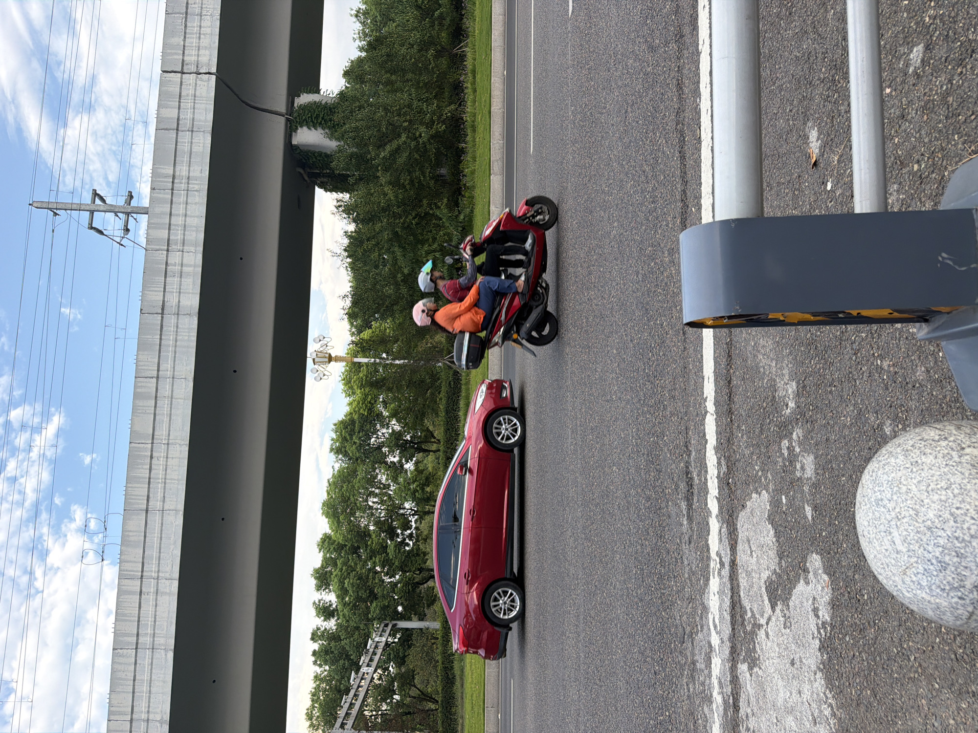 On a clear day, a red sedan is parked on the right side of a paved road. Behind the sedan, a red scooter is traveling away from the viewer. The scooter is carrying two individuals, both wearing helmets. The person in front is wearing a red shirt and blue jeans, while the person behind is wearing an orange jacket and blue jeans. The scooter has a black storage box mounted on the back. In the background, there is a large, elevated concrete structure, possibly a bridge or overpass, with power lines running above it. Green trees and vegetation line the side of the road. The scene appears to be in Jiangjin, China.