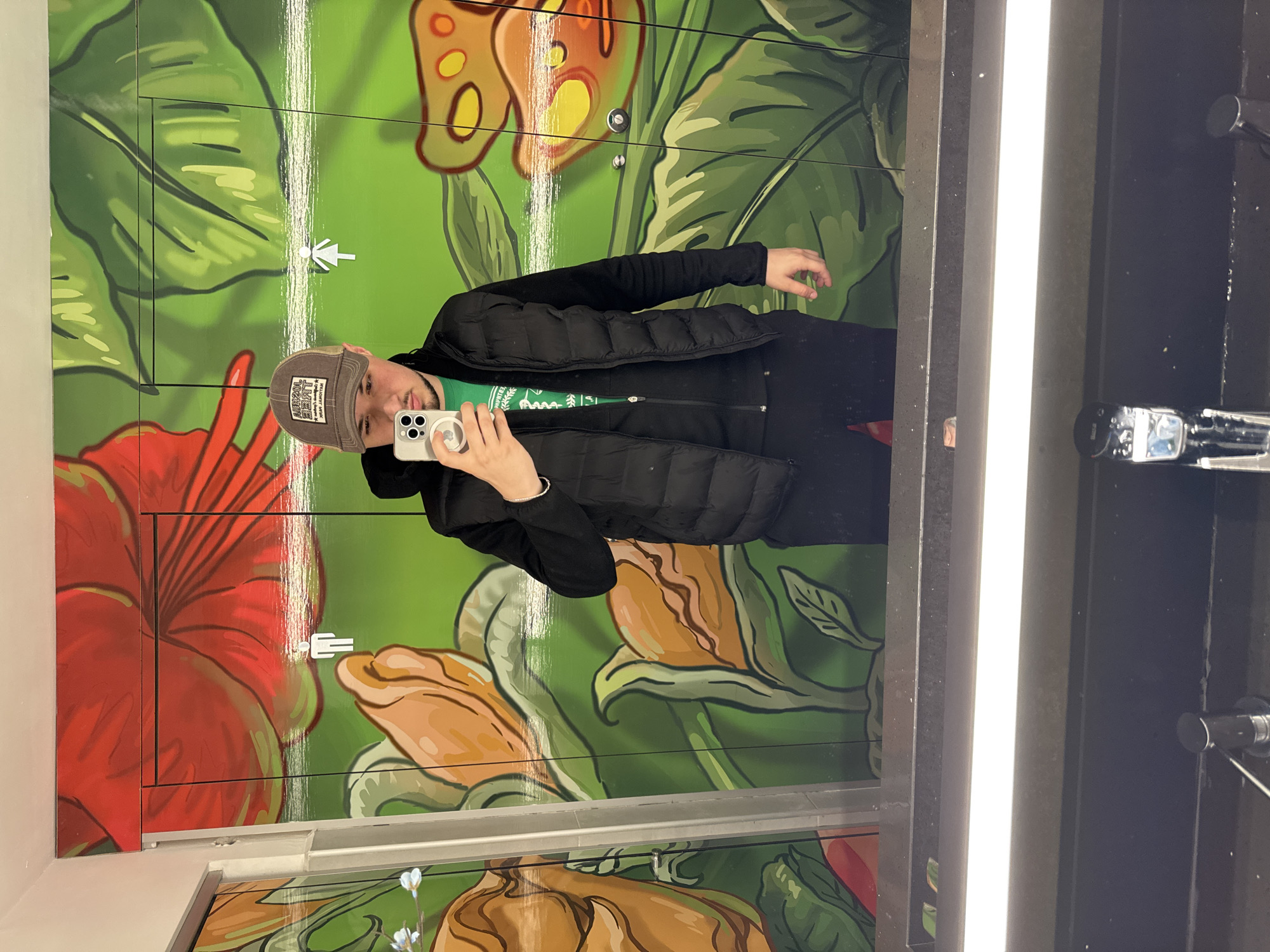 A man is taking a selfie in a restroom in Varna, Bulgaria. He is wearing a gray baseball cap, a black jacket, and a green shirt. He is holding a white smartphone. The restroom has a brightly colored floral mural. Restroom signs are visible on the mural. A light fixture is visible above the mirror.