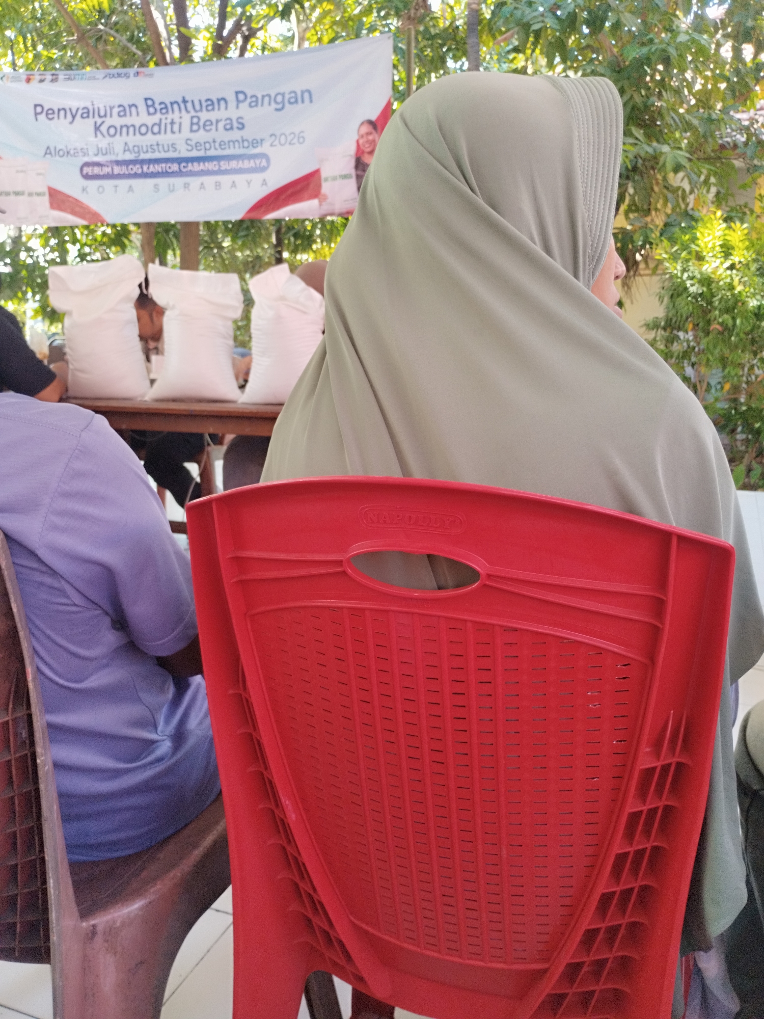 The image depicts an outdoor scene with a banner in the background that reads "Penyaluran Bantuan Pangan Komoditi Beras Alokasi Juli, Agustus, September 2026 PERUM BULOG KANTOR CABANG SURABAYA KOTA SURABAYA." This translates to "Rice Commodity Food Aid Distribution July, August, September 2026 PERUM BULOG SURABAYA BRANCH OFFICE CITY OF SURABAYA." In the foreground, a person wearing a purple shirt is seated, facing away from the camera. To the right, another person, partially obscured by a red chair, is wearing a light green hijab and looking to the right. Three large white sacks are visible behind them, placed on a wooden surface. The environment appears to be sunny, with trees and foliage in the background. The red chair in the foreground has the word "NAPOLY" embossed on it.