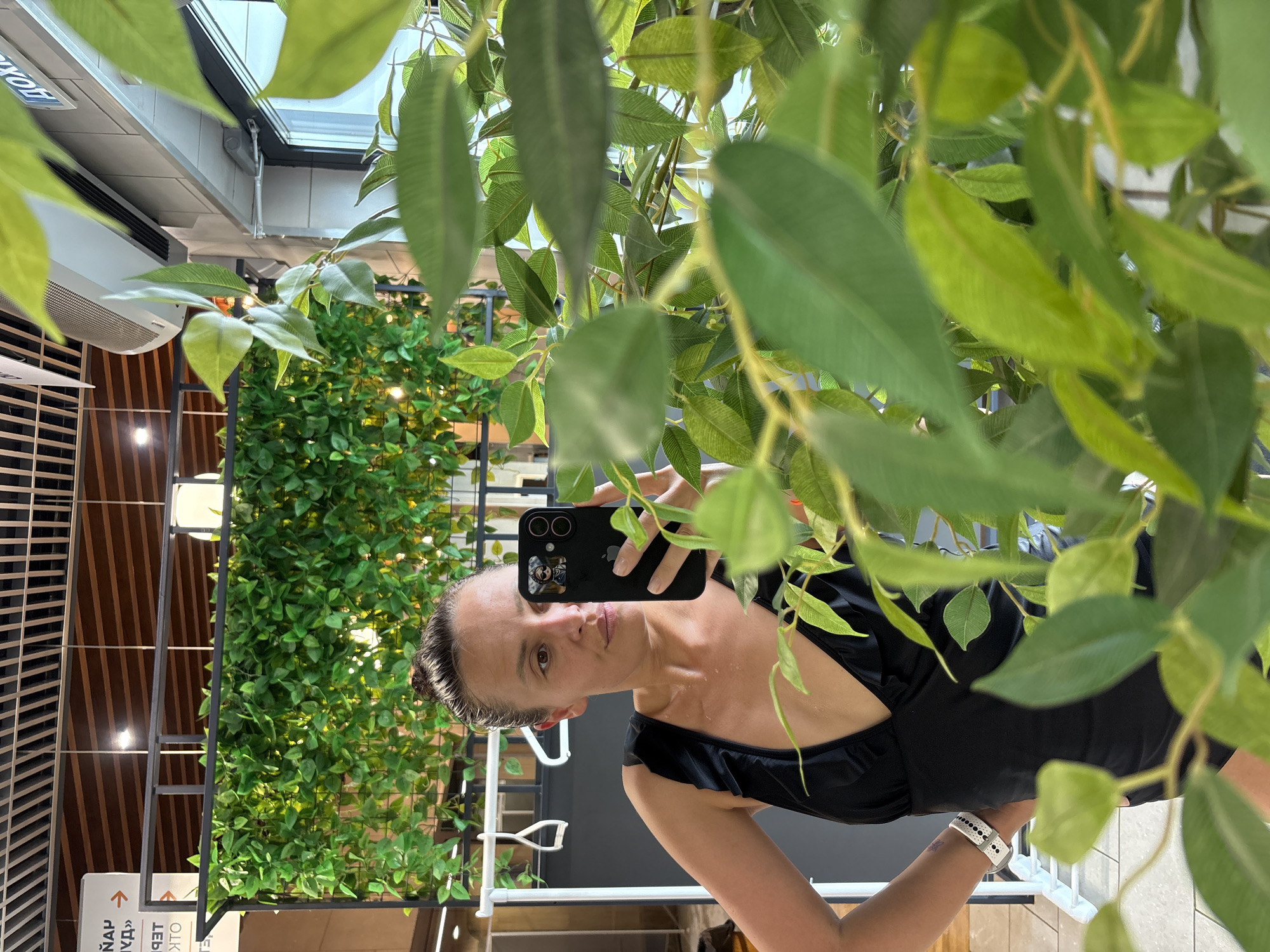 A woman is taking a selfie with a black smartphone in what appears to be an indoor establishment with greenery. The woman is wearing a black dress with ruffles and a white smartwatch. She is positioned amongst green leafy plants, some of which are blurred in the foreground. Behind her, there is more artificial foliage, possibly a green wall. The setting has overhead lighting, and structural elements such as metal frames and ceiling fixtures are visible. A sign with text is partially visible. The location is in the city of Votkinsk, Russia.