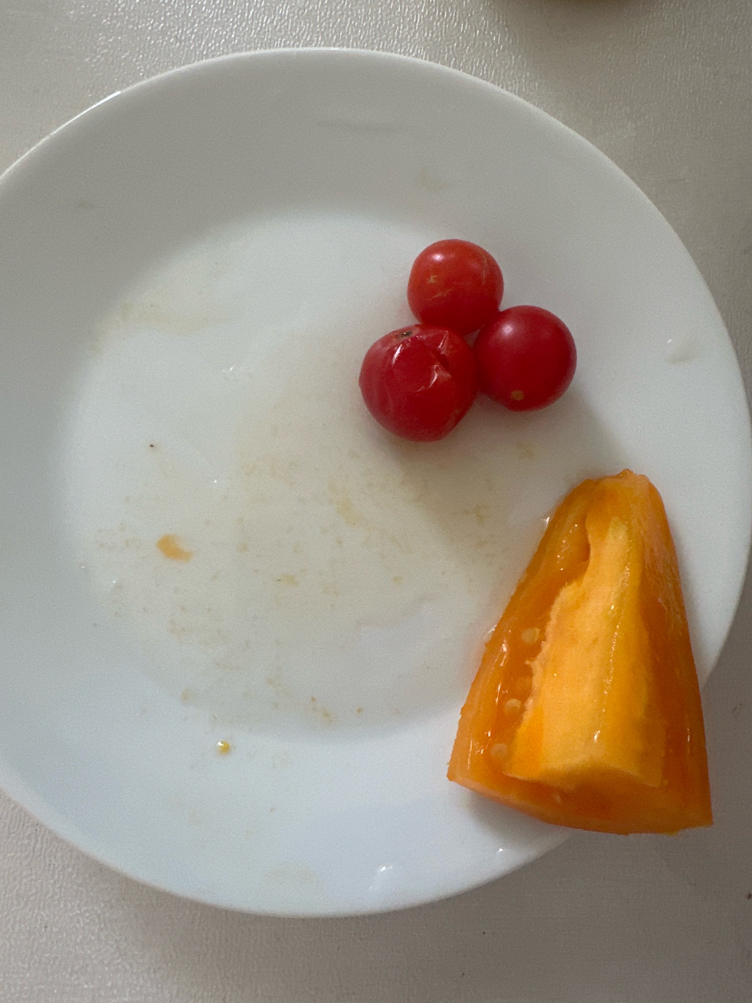 Three cherry tomatoes and a slice of orange bell pepper are on a white plate, which is on a light-colored, textured surface. The plate has some residual moisture and small food particles. The scene is an indoor, domestic setting. There is no visible indication of the city of Nevinnomyssk, Russia.