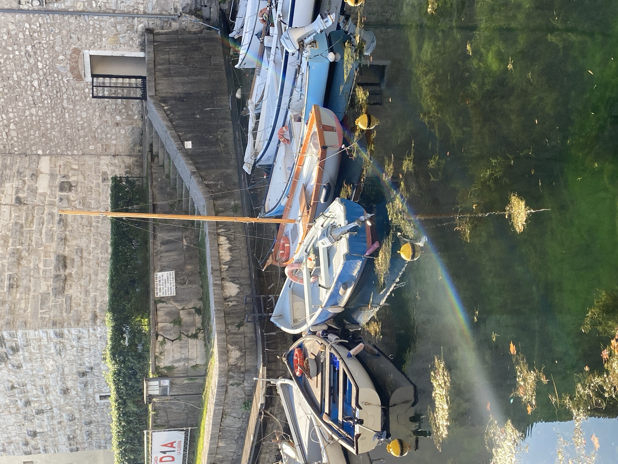 This image depicts a waterfront scene in Riva del Garda, Italy. Several small boats are moored in the clear, green water, some with their masts erect. The boats are of various colors, including white, blue, and orange. Yellow buoys float in the water, and a faint rainbow is visible, reflecting off the surface. In the background, a stone building with a grassy area and stairs leading up to a doorway is visible. The sun is shining, casting shadows and highlighting the textures of the stone. There are no people visible in the image. The overall impression is one of a peaceful, sunny day in a picturesque Italian town.