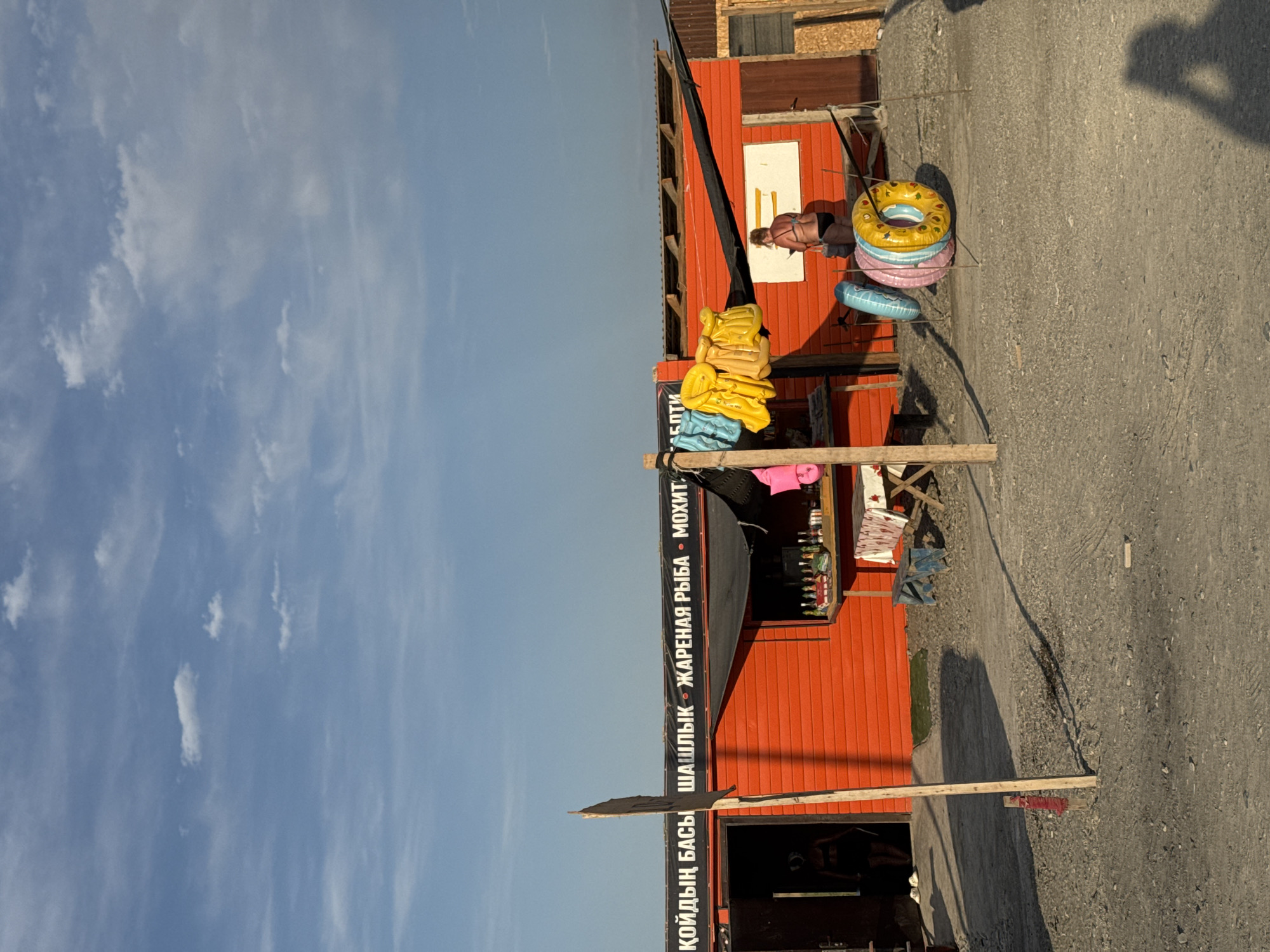 The image depicts an outdoor scene at a beachside stall in Zharbulak, Kazakhstan. The stall is painted bright orange and features a black awning. Above the awning, white text lists various offerings: "КОЙДЫҢ БАСЫ", "ШАШЛЫК", "ЖАРЕНАЯ РЫБА", and "МОХИТ".
In front of the stall, several inflatable rings are stacked and propped up with sticks. To the left, a doorway leads into the stall, where a person is partially visible. To the right, a woman in a bikini stands by the counter of the stall, seemingly interacting with the items displayed there. A table with a patterned tablecloth is set up in front of the stall, with bottles and what appear to be drinks or food items arranged on it. A small blue bench is situated near the table.
The ground is covered in gravel and dirt, with shadows cast by the stall, objects, and people. The sky above is blue with scattered clouds. A wooden post stands prominently in the foreground, partially obscuring the view of the stall. The overall impression is that of a casual, possibly tourist-oriented, establishment in a sunny outdoor environment.