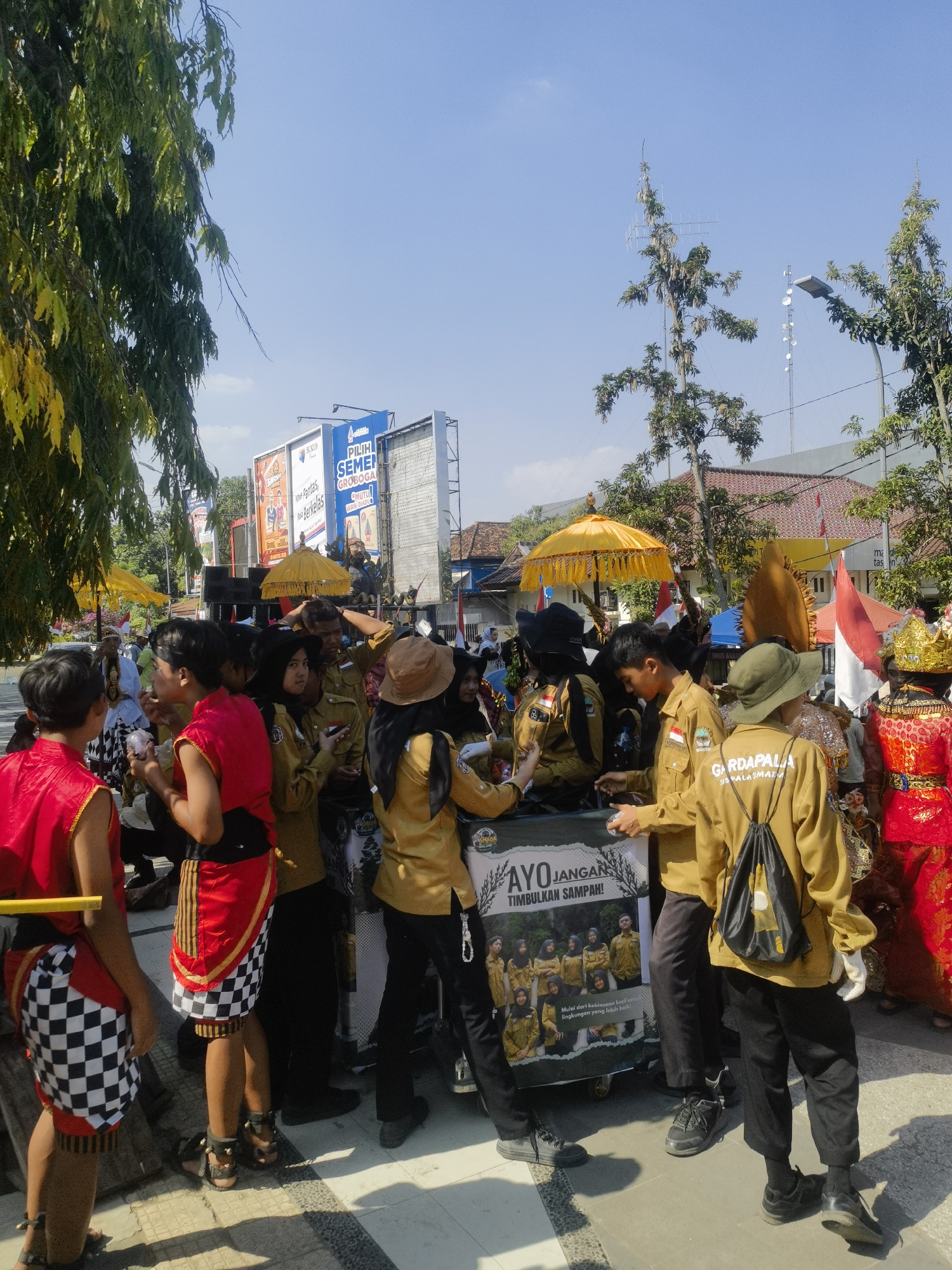 This image captures a lively street scene in Blora, Indonesia, likely during a cultural event or parade. A group of people, many wearing distinctive tan uniforms with black accents and some adorned in traditional red and black attire, are gathered around a cart. The cart displays a banner with Indonesian text that reads "AYO JANGAN TIMBULKAN SAMPAH!" (Let's not create trash!), accompanied by a photo of people in similar uniforms.
Several individuals are interacting, with some looking at the cart and others conversing. One person in the foreground wears a red vest over a black shirt and a patterned sarong, while another in a similar red outfit has a checkered pattern on their lower garment. The individuals in tan uniforms appear to be part of an organization or school, as indicated by patches on their clothing. One person is wearing a green hat and another a brown hat.
In the background, billboards are visible, including one advertising "SULAIH SEMEN GROBOGAN". Trees with green and yellowing leaves line the street, and a bright yellow umbrella is prominently displayed, suggesting it might be a decorative element of the event. The sky is clear and blue, indicating a sunny day. The ground appears to be paved with tiles or concrete. The overall atmosphere suggests a public gathering with an emphasis on environmental awareness and local culture.